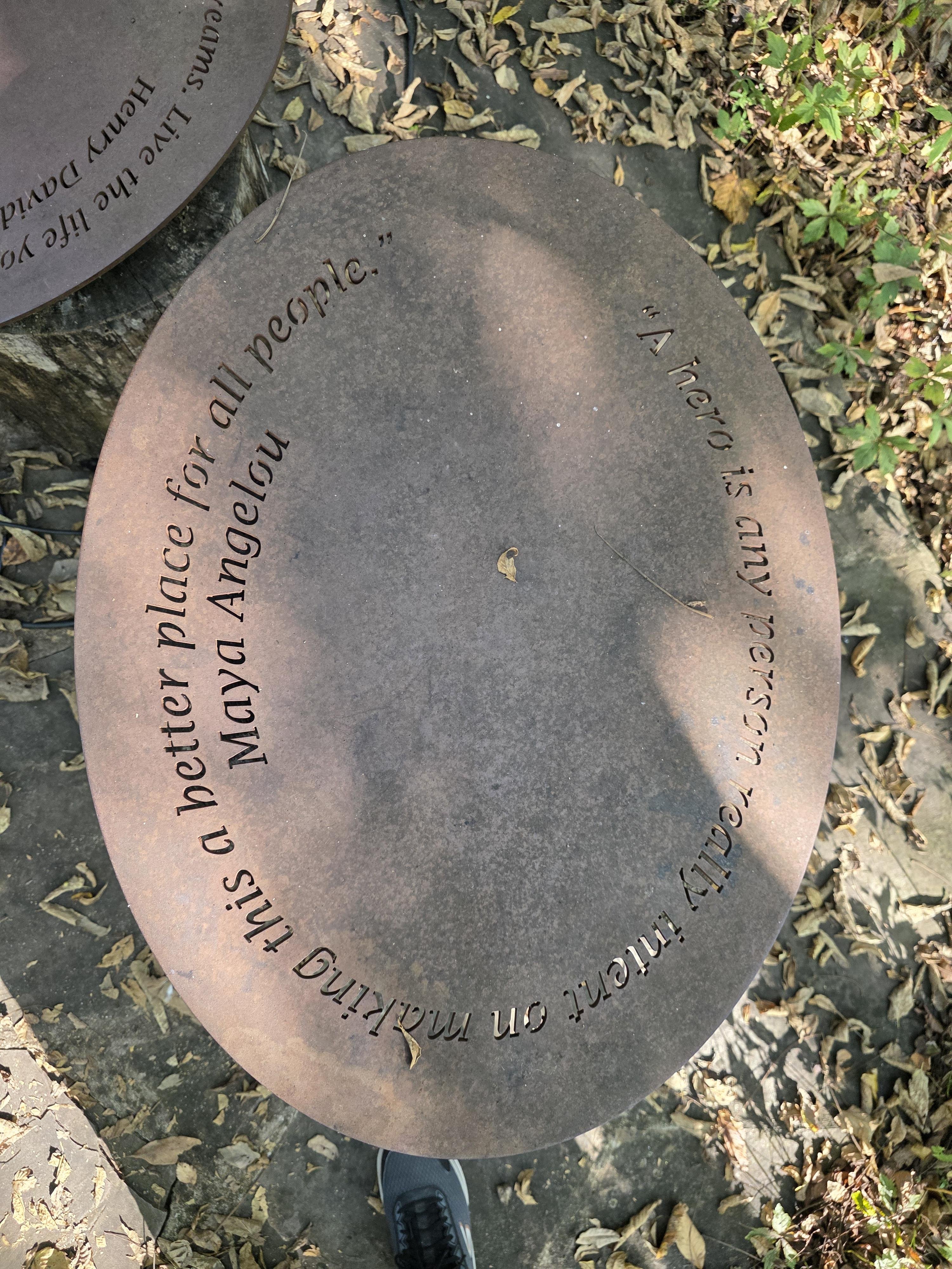 Oval-shaped metal plaque set on a tree stump, inscribed with the quote, “A hero is any person really intent on making this a better place for all people.” Signed with the name Maya Angelou. Surrounding area features fallen leaves and a glimpse of a foot wearing a black sneaker.