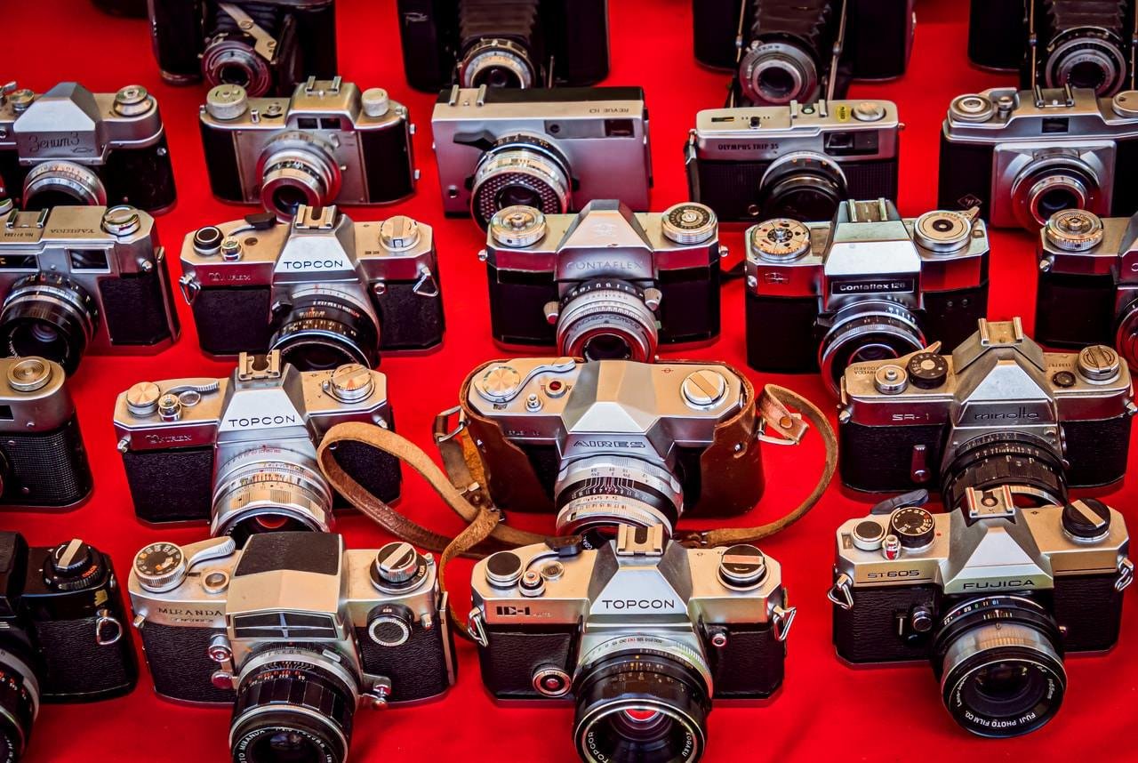 The image shows a collection of vintage cameras displayed on a red surface. There are various models and brands, with some cameras featuring leather straps. It's a visually appealing assortment of retro photographic equipment.