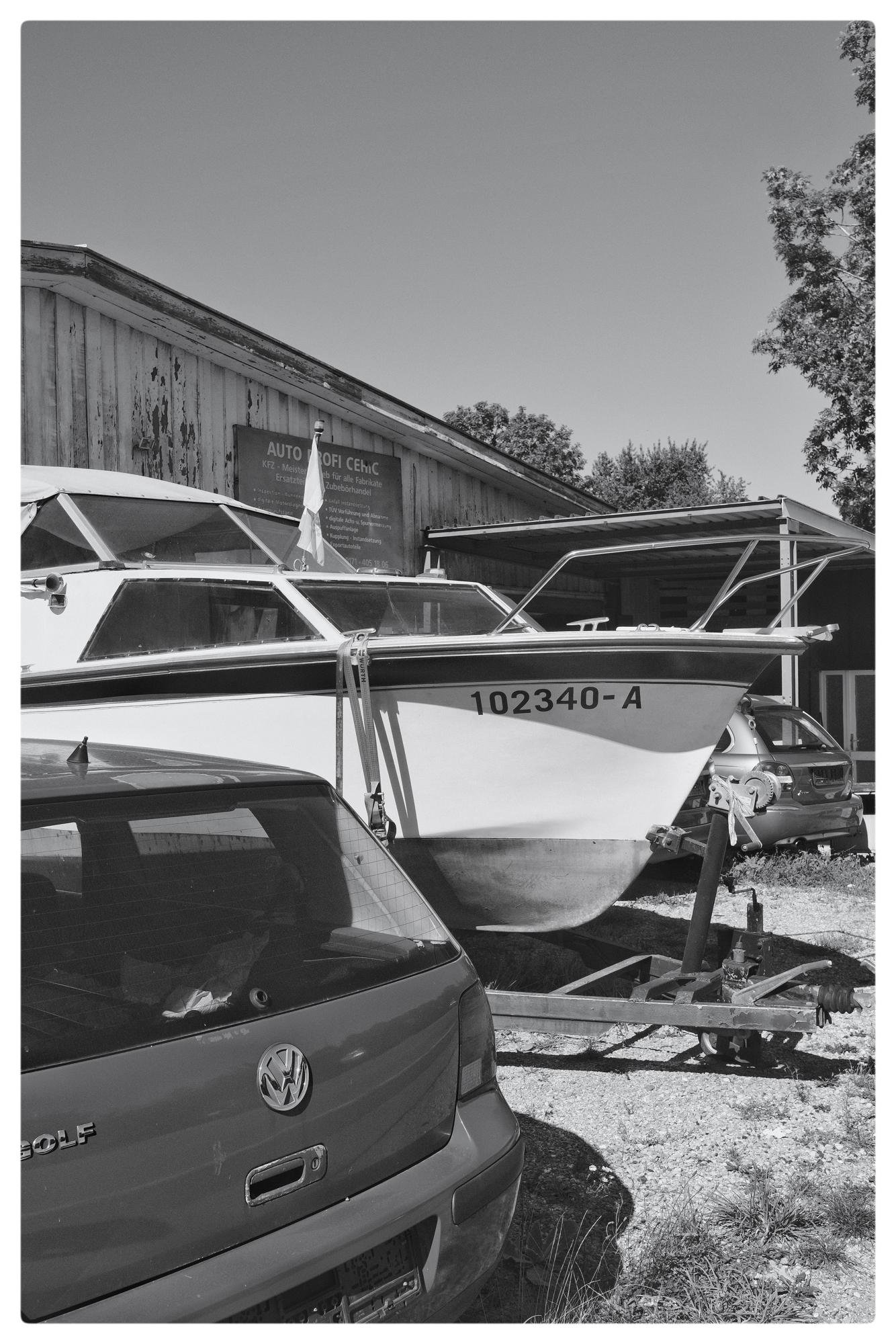 Black and white image of a boat with the number 102340-A on a trailer parked next to a Volkswagen Golf. In the background, a wooden building with a sign reading "AUTOPROFI CEHIC" is visible. Other vehicles and trees are in the background, and the scene is set on gravel with clear skies above.