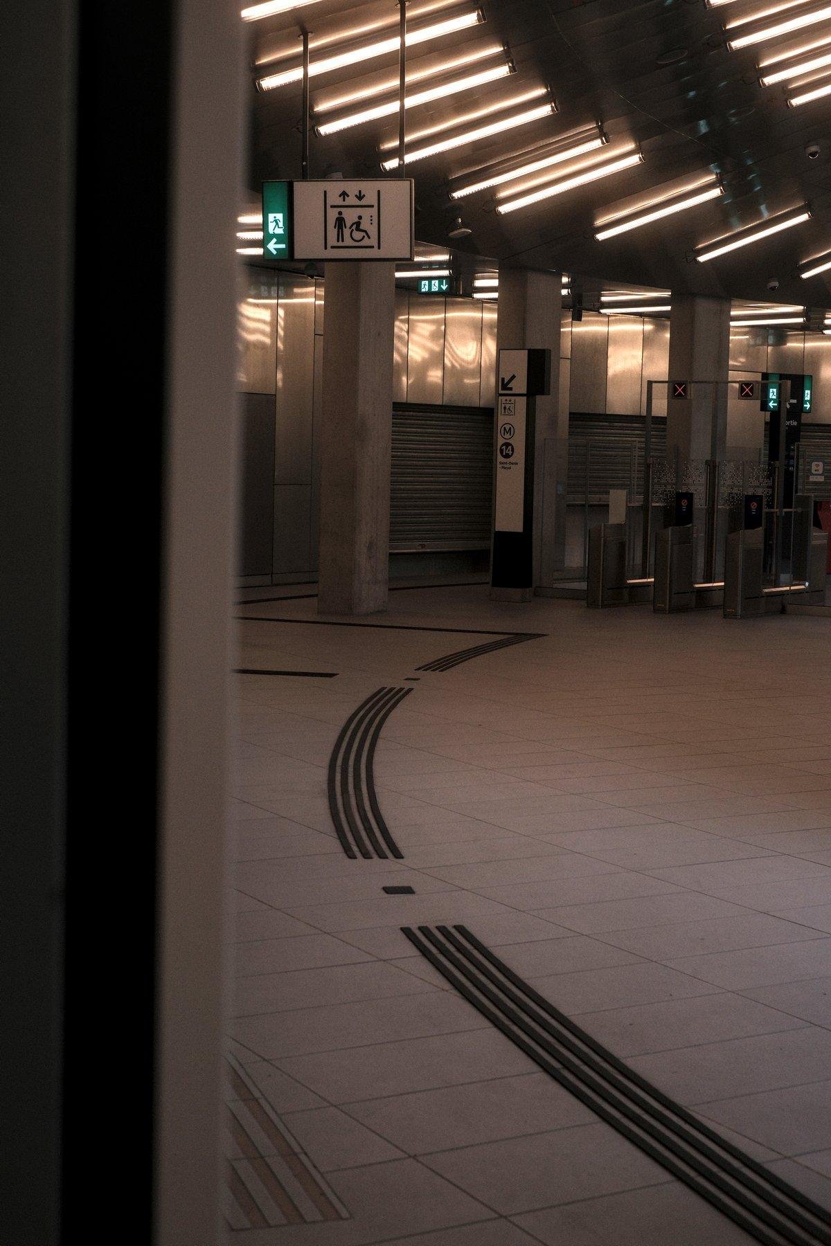 Photo verticale. Un couloir de métro courbé vers la droite, éclairé par des néons posés en suivant la courbe et menant à des bornes d’accès côté sortie à en croire les croix rouges allumées. Au sol, un guide en relief pour malvoyants qui suit la courbe aussi et tourne à 90° à gauche avant les sorties
