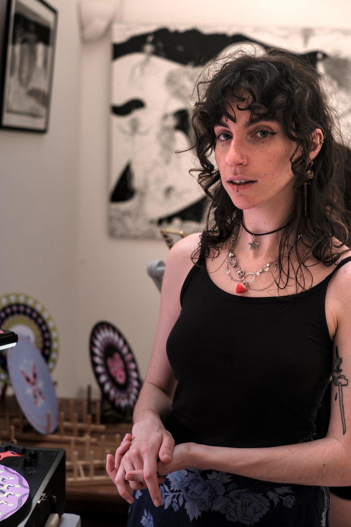 Photo verticale. Portrait d’une jeune femme brune au cheveux ondulés, en débardeur noir et jupe longue noire aux motif végétaux bleu. elle est entrain de m’expliquer son œuvre d’art pendant que capture des images