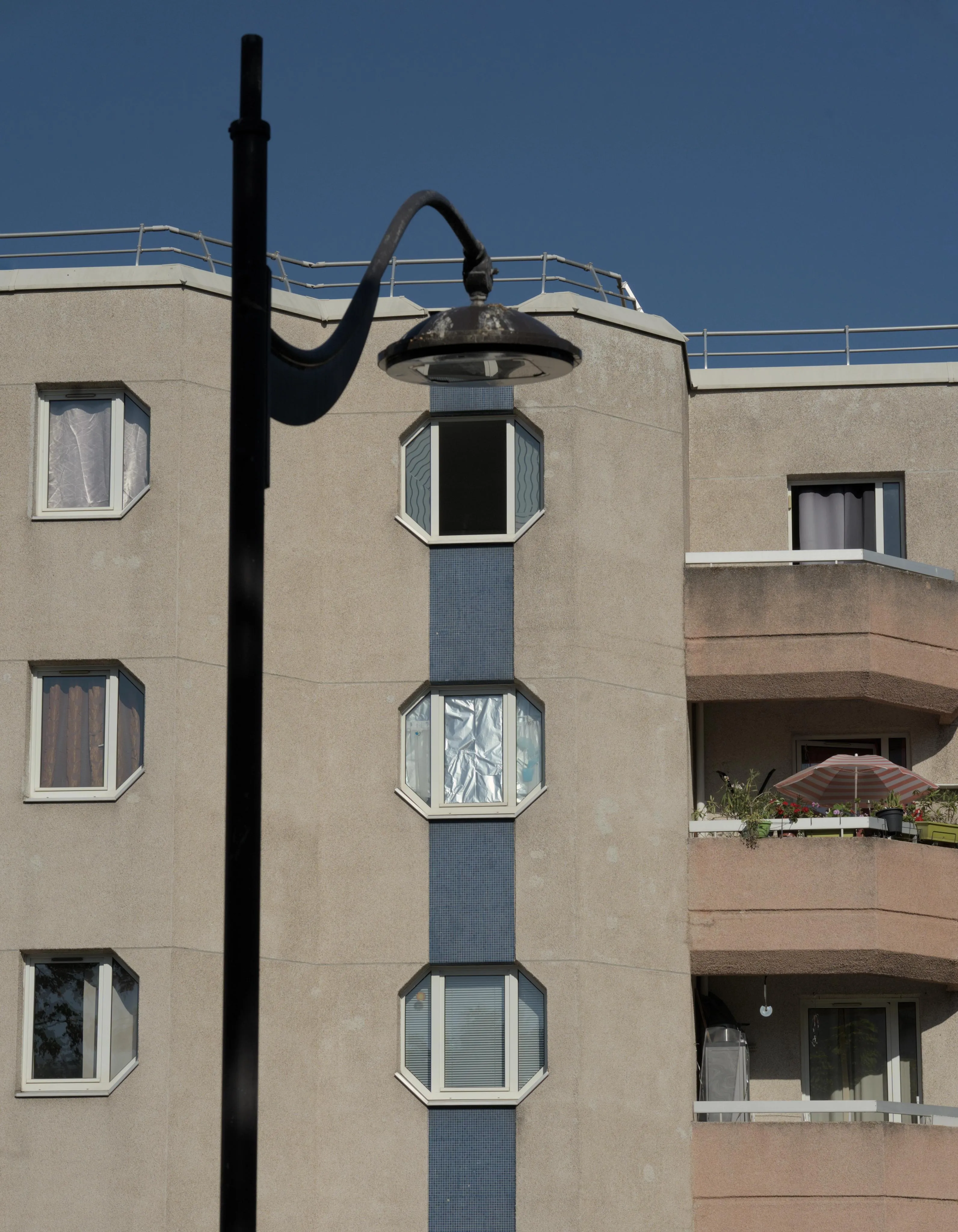 Photograph of a black street lamp post in front of an habitation building.  The perspective puts the street light lamp at the top of a column of windows on the building. The column of 3 windows has a blue tiling decoration while the rest of the building facade has a light color. The inside of the middle window is covered with aluminum foil, reflecting the light from outside. On the left of the lamp post pole, another column of 3 windows. On the right hand side there are balconies at each floor. The middle balcony has plants and a parasol. Over the building, a clear blue sky covers the top of the image.