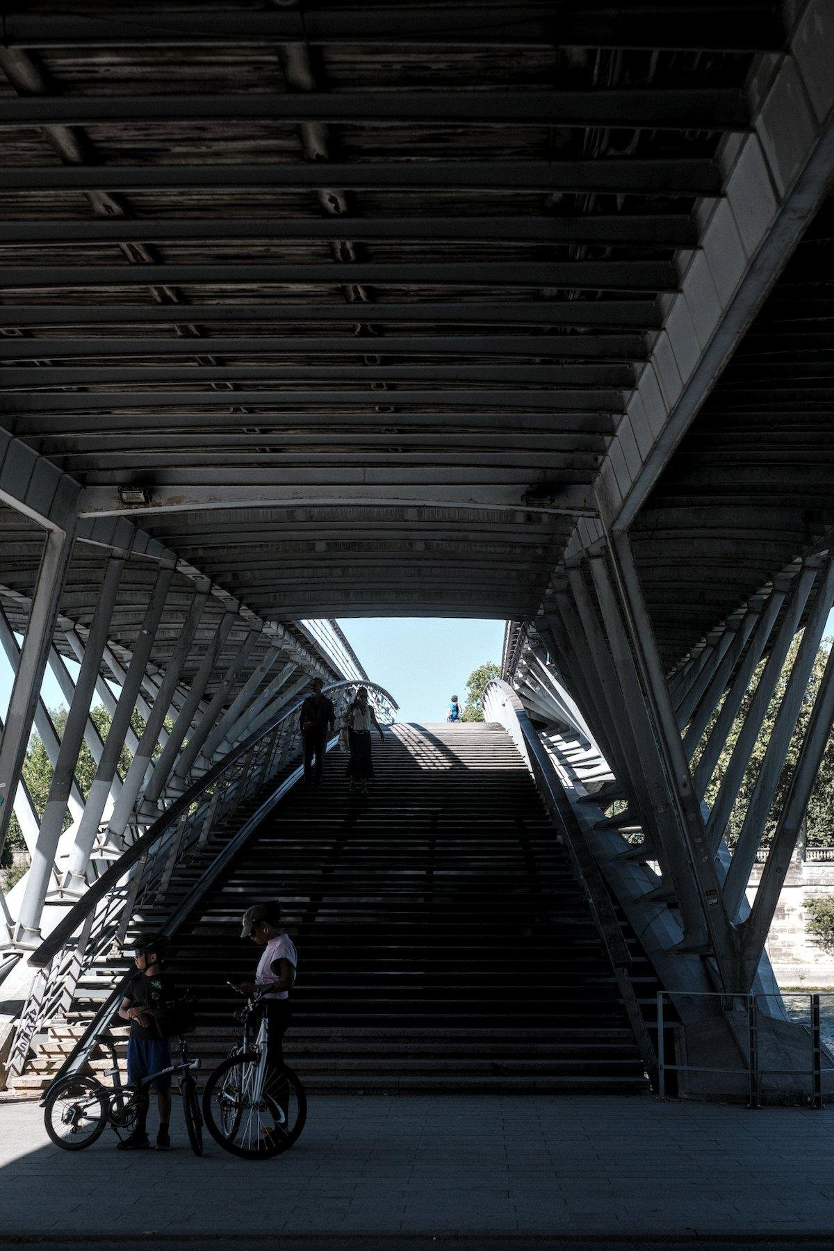 Photo verticale. Sous un pont en acier, un vaste escalier permet de choisir de passer dessus ou aller dessous pour rejoindre les quais. Au premier plan 2 personnes à côté d’un vélo de profil, 2 personnes sont sur la gauche de l’escalier et le descendent, du ciel bleu est visible au bout de l’escalier en arrière plan