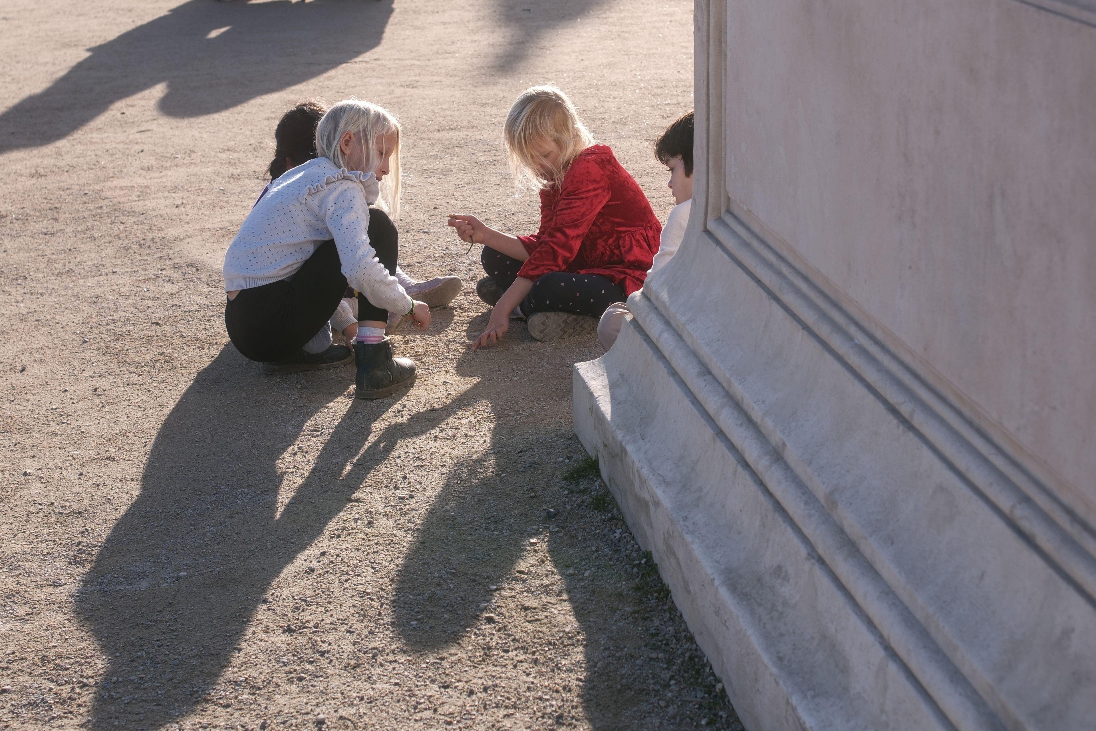 Photo horizontale. Un groupe d’enfants accroupis ou assis par terre jouant à dessiner sur le sol sablonneux. À droite, un bout de socle en marbre blanc.