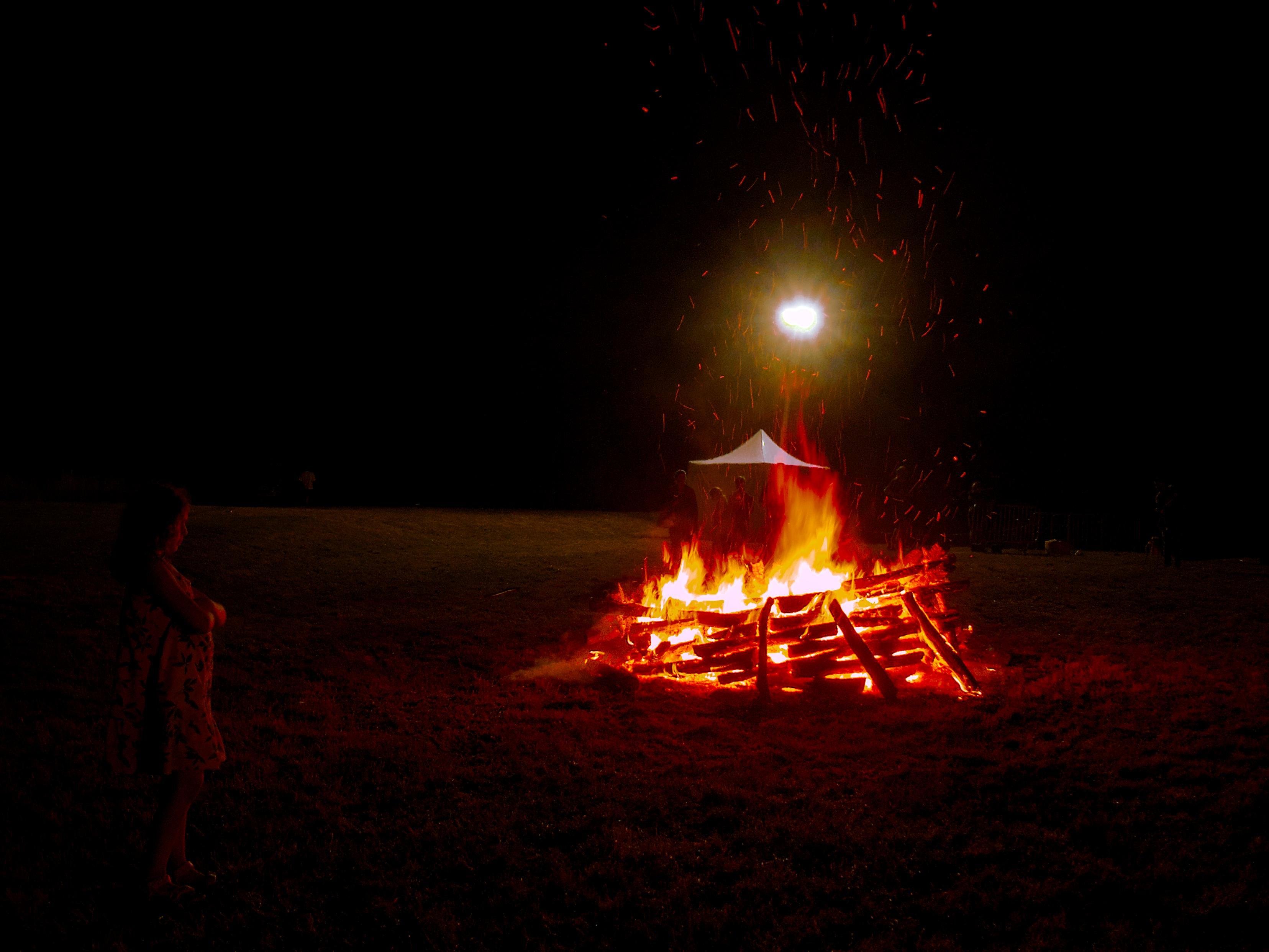 Bonfire built from same sized and well structured wood logs burning in an open field at night, with sparks rising; a bright light above a small white tent behind the fire and a silhouetted profile of a person looking at the fire, arms crossed, at the bottom left corner.