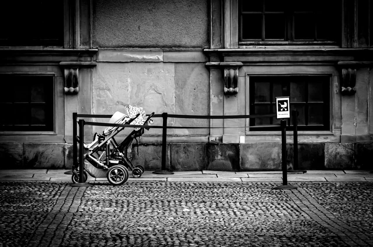 Black and white image depicting an empty baby stroller with a patterned blanket, parked against a historic building facade. A rope barrier runs parallel to the stroller, featuring a sign with indistinct symbols. The ground is paved with cobblestones, and the building has two windows framed by ornate stonework.