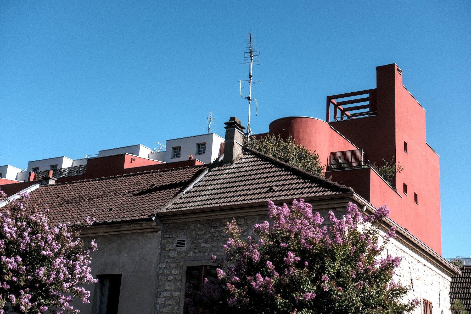 Photo horizontale. Derrière des toitures marron, classique un bâtiment moderne rouge ressort sur le ciel bleu