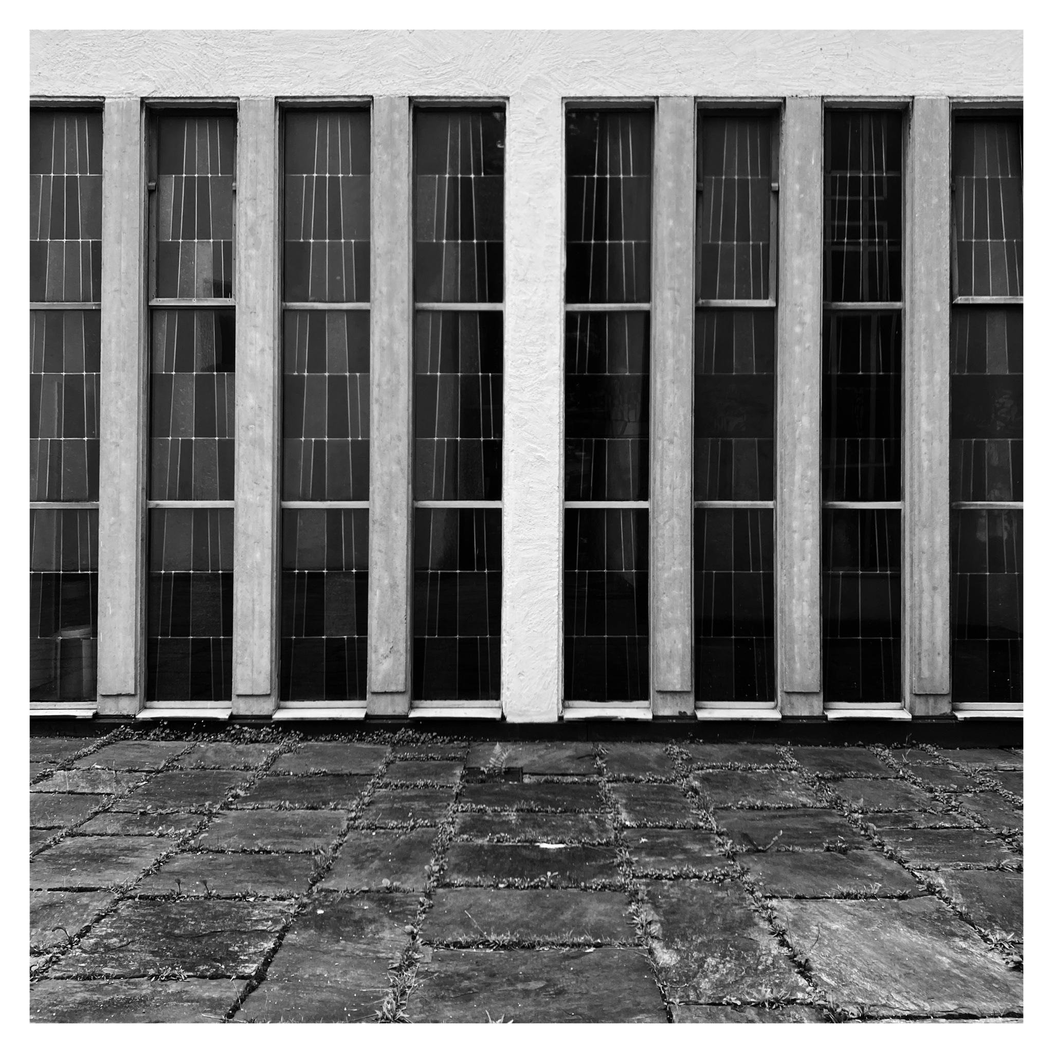 Black-and-white photo of a building facade with two rows of tall, narrow windows separated by thick concrete vertical columns, above a paved stone walkway in the foreground.