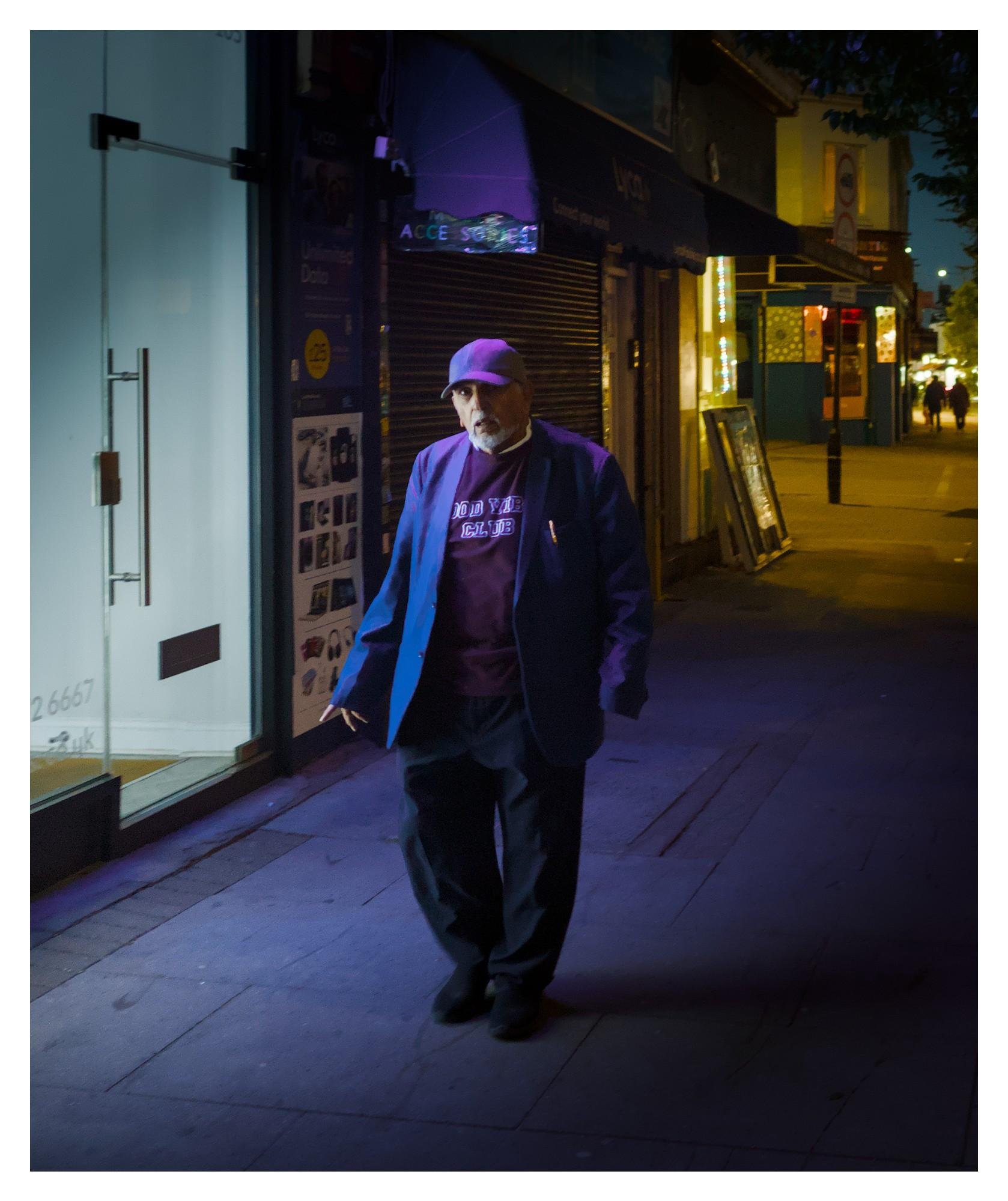 Older man in a cap and jacket walking alone on a nighttime sidewalk, lit by blue and purple streetlight; closed shop shutters and a glass storefront line the street, with illuminated signs and distant pedestrians in the background.