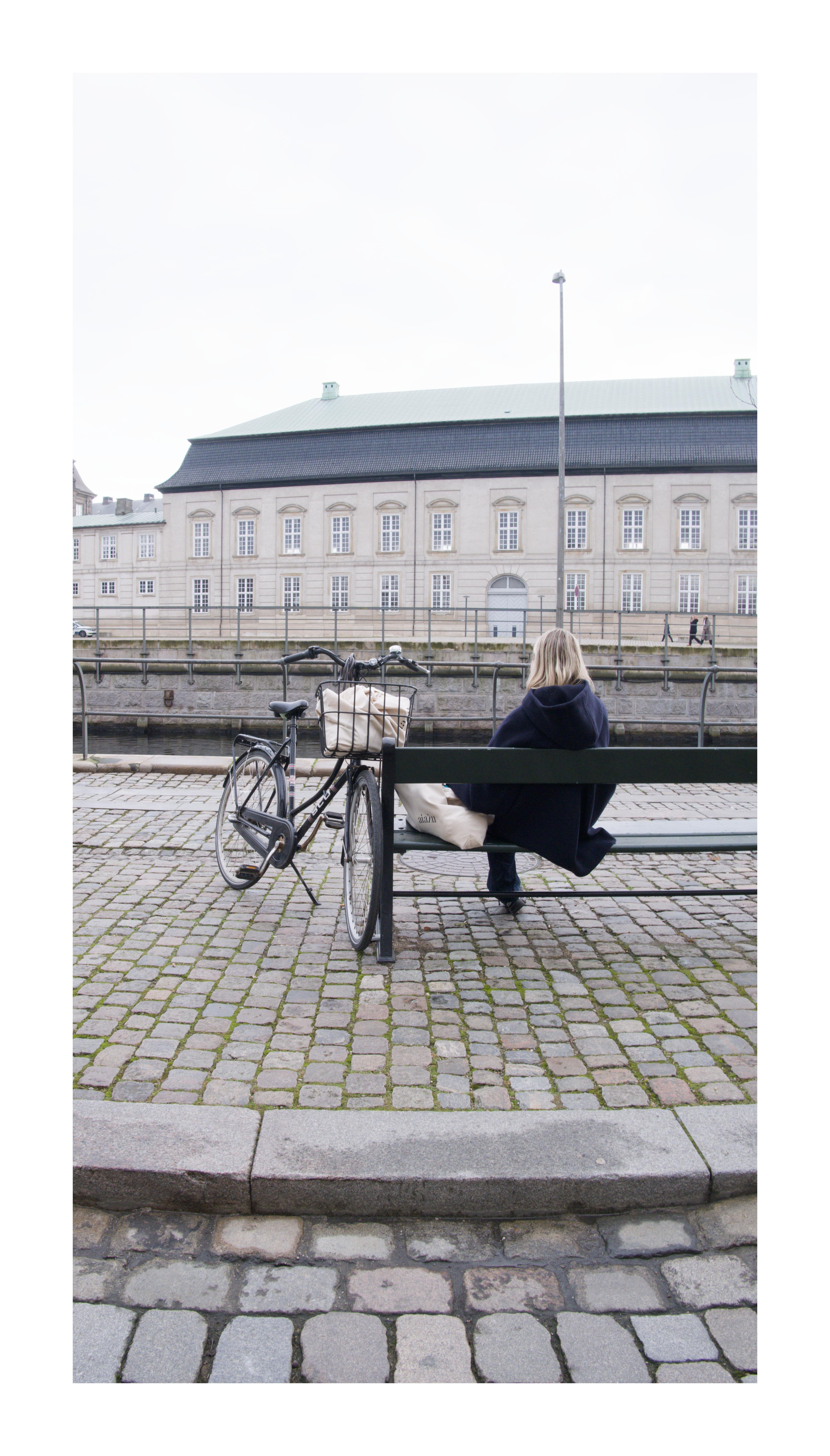 Person with blond hair sitting on a green bench beside a parked bicycle with a front basket on a cobblestone walkway next to a canal; across the water is a long, pale building with many windows and a tall lamppost.