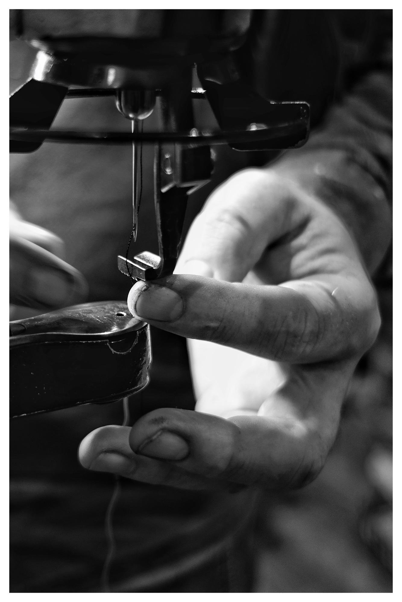 Close-up black-and-white photo of a sewing machine needle and presser foot, with hands guiding fabric under the needle; thread visible hanging below the machine.