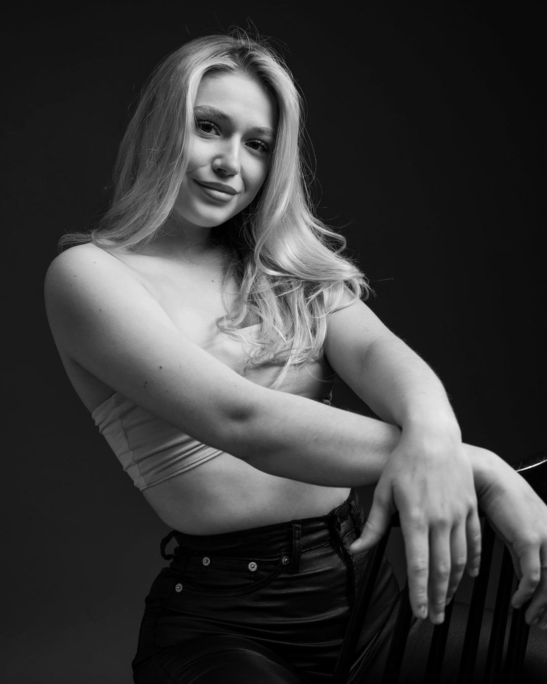 The image is a black and white portrait of a person with long hair sitting in a relaxed pose. They are wearing a sleeveless top and dark pants, with their arms resting on the back of a chair. The background is plain and dark, emphasizing the subject.