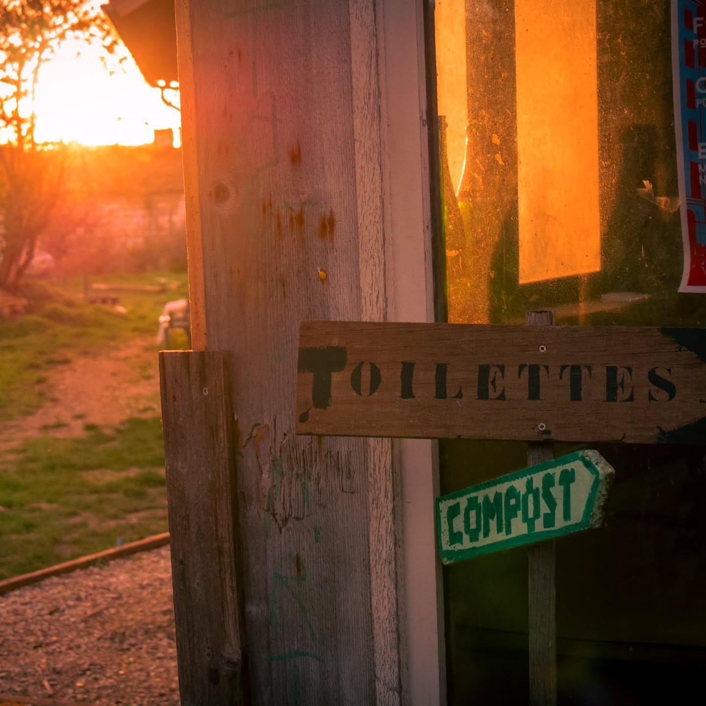 The image shows a wooden structure with two signs. One sign reads "TOILETTES" (French for "Toilets") and the other says "COMPOST," with an arrow pointing in a direction. The setting appears to be outdoors, with grass and a pathway visible, and the image is taken at sunset or sunrise, creating a warm, golden glow.