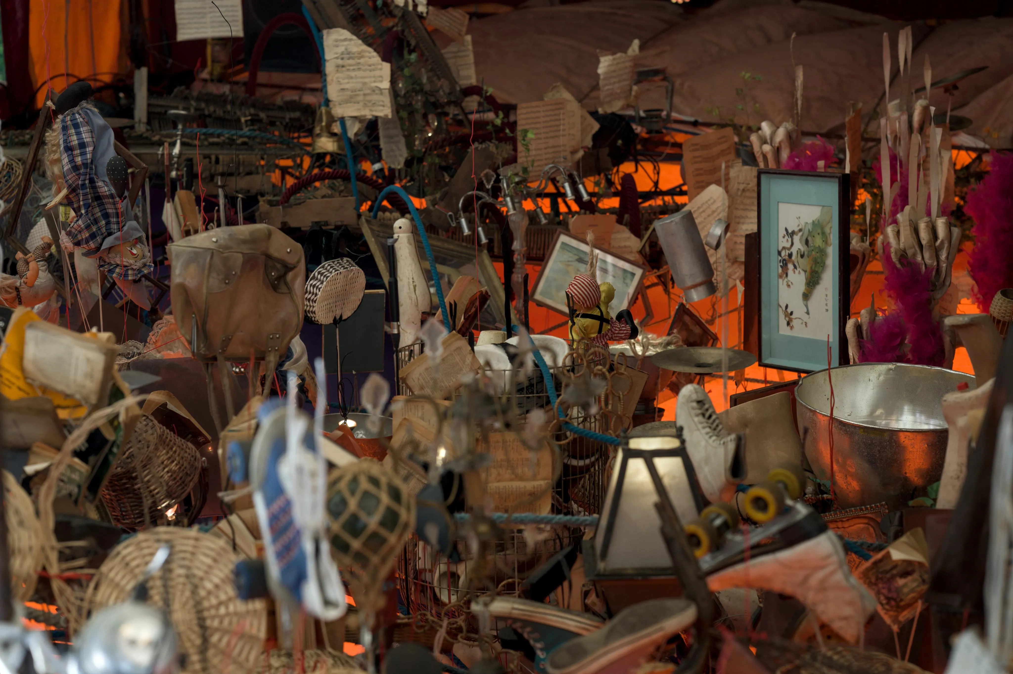 Photograph of various used household objects appearing to float, tethered to the bottom of image. The various objects include framed paintings, used shoes, kitchen utilities, old handbags, music sheets, dolls.