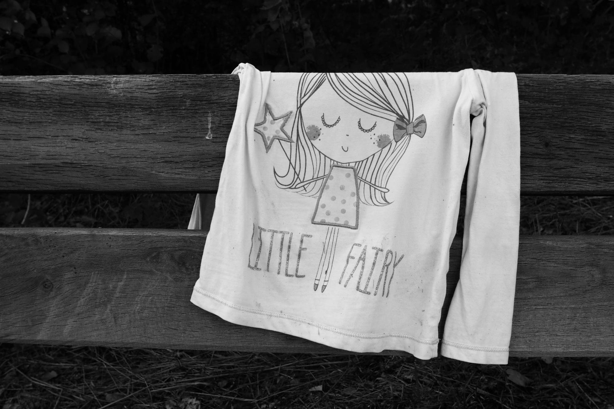 Black-and-white photo of a child’s long-sleeve shirt draped over a wooden bench, featuring a cartoon fairy holding a star wand and the text “LITTLE FAIRY.”