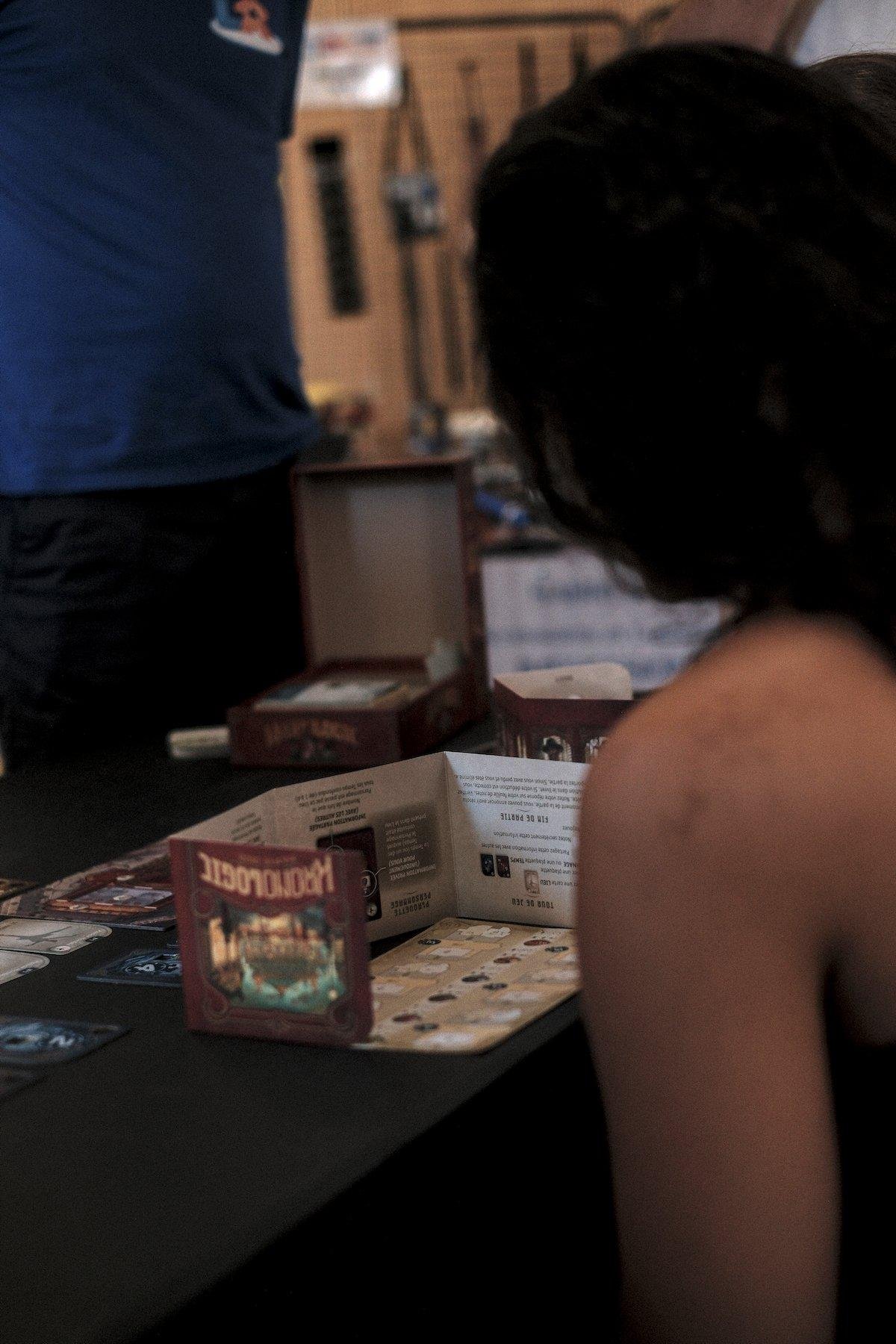 Photo verticale. Une table avec une nappe noire avec une partie de jeu de société en cours (Kronologic). Une jeune femme dont on ne voit que l’épaule et les cheveux noirs est devant sont plateau personnel de jeu, caché par un écran qui est posé à l’envers, comme en témoigne le nom du jeu à l’envers