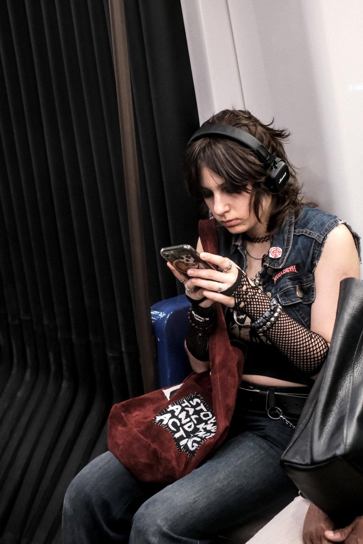 Photo verticale. Une jeune femme (ado ?) assise dans le métro près des «&nbsp;accordéons&nbsp;» de jonction entre wagons, brune cheveux courts un peu en bataille, casque audio sur la tête de marque marshall, veste en denim bleu foncé sans manche, mitaine fishnet, jean, sac baudruche bordeaux, entrain de taper sur son tél