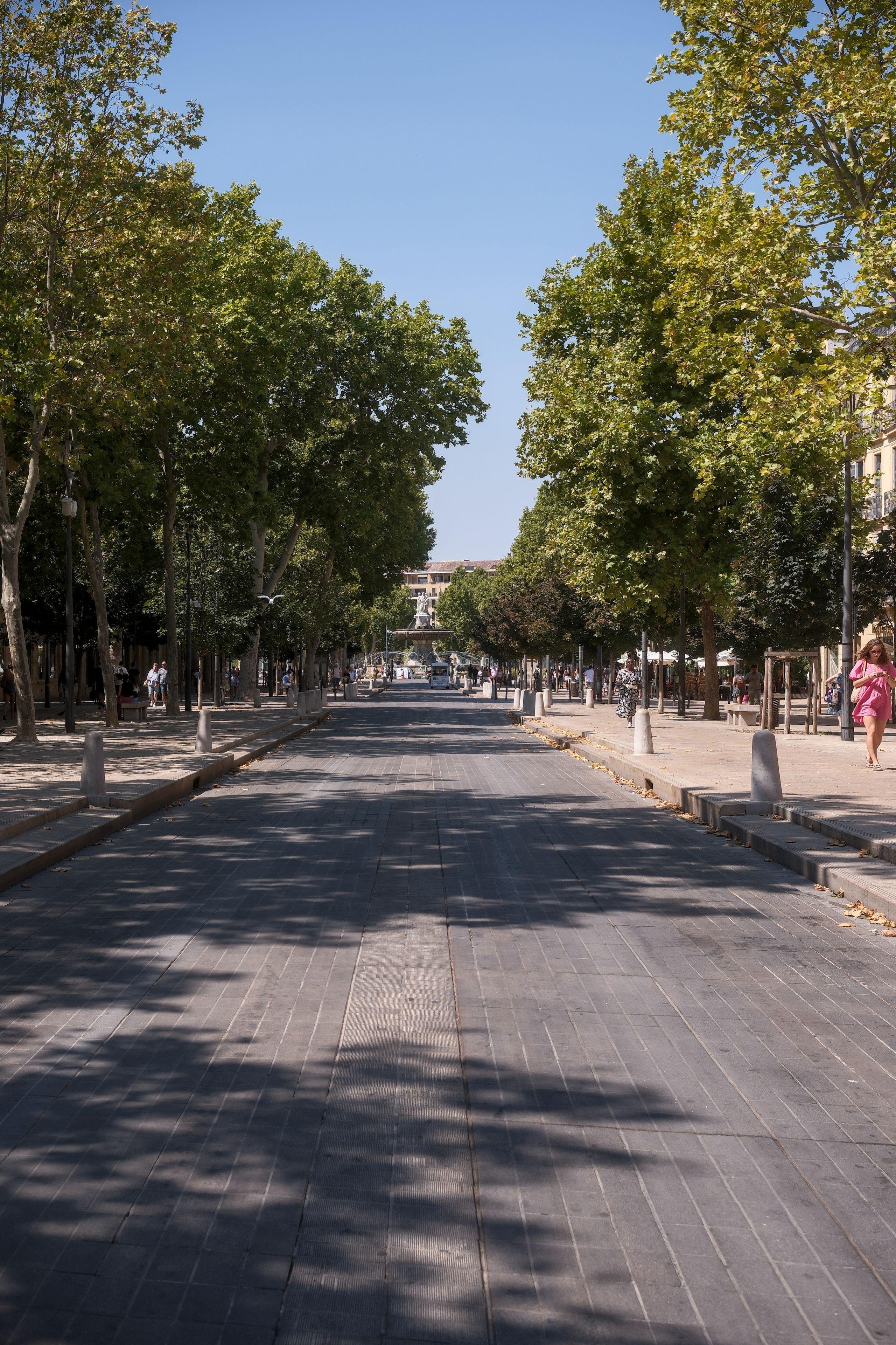 Photo verticale. Une avenue pavée moderne, vide de voitures (un minibus au fond à peine visible). Des piétons épars sur chaque côté