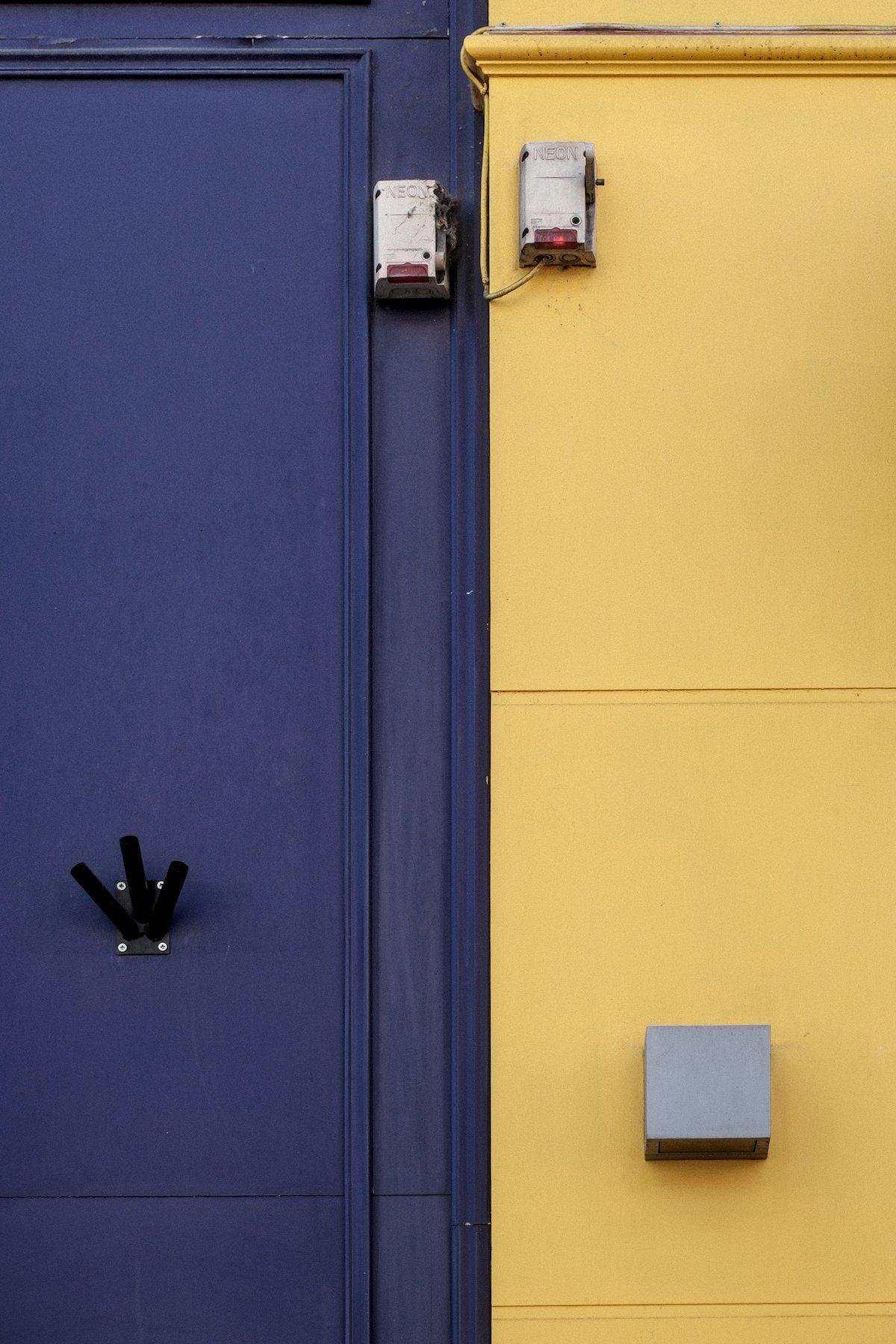 Photo verticale. Mur extérieur de 2 boutiques différentes, chacune ayant peint son mur d’une couleur différente avec délimitation nette au centre de l’image. Violet à gauche, jaune à droite