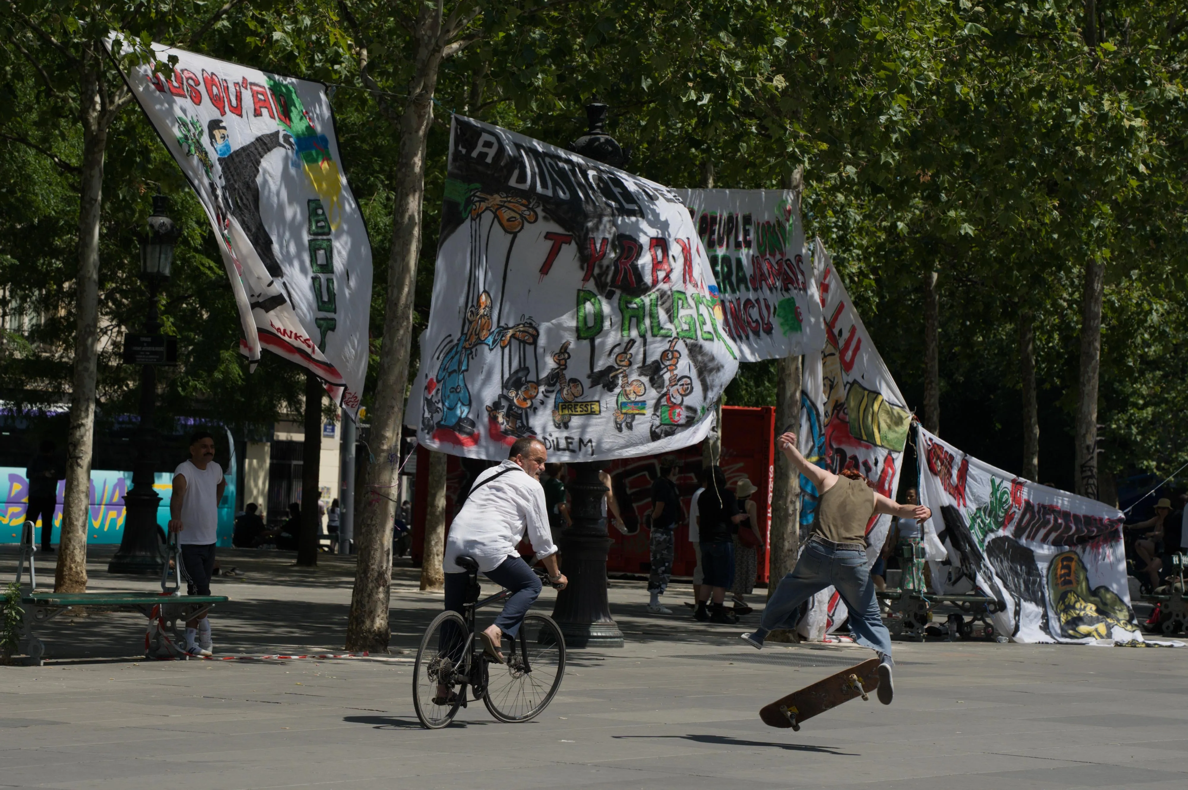Photograph of a skater doing a flip trick on a public square bordered with trees. The skater is facing away from the camera. The skater's feet are in the air above the skateboard, their legs are half extended and their arms are extended to their back. A cyclist riding besides the skater is looking at the skater. In the shade of the trees, another man is watching the skater, his arm resting on a public bench. In the background between the trees, large banners are suspended overhead with paintings and writings denouncing social injustice.