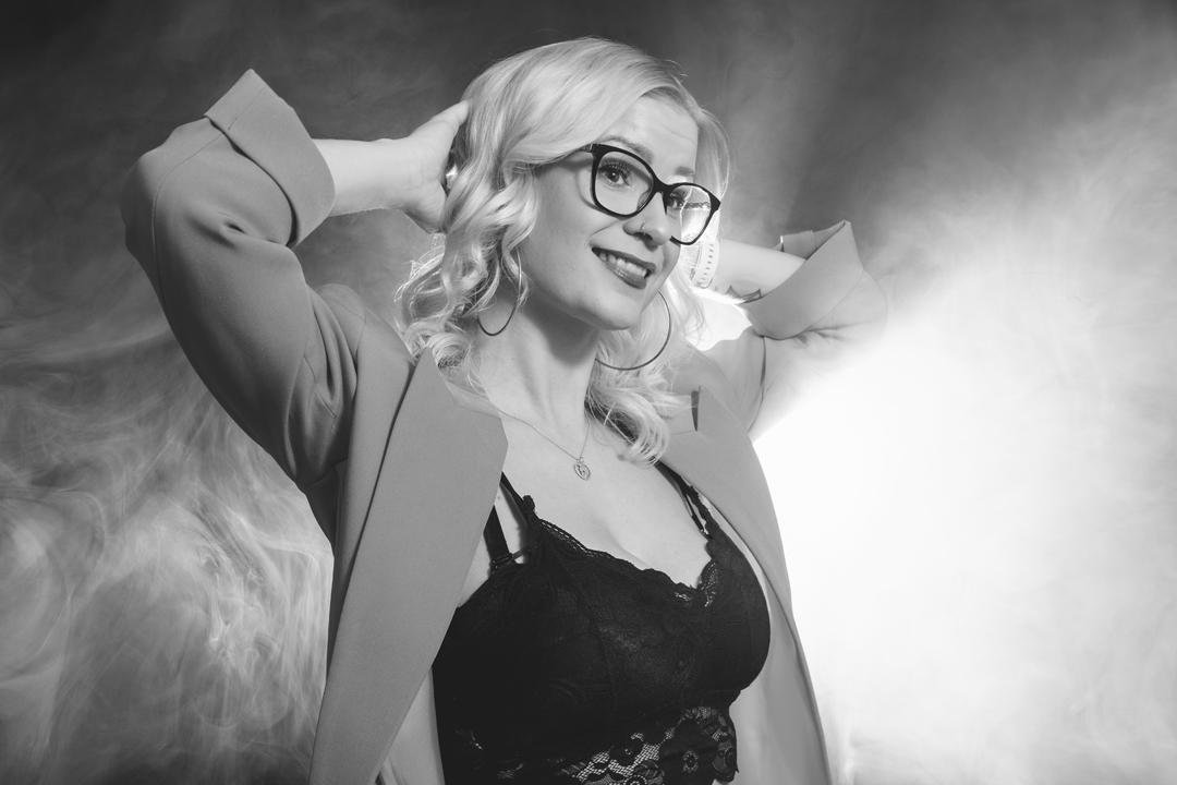 This is a black and white photo of a person wearing glasses and a lace camisole under a jacket. The individual has light hair styled in waves and is posing with hands behind their head against a smoky or misty background.