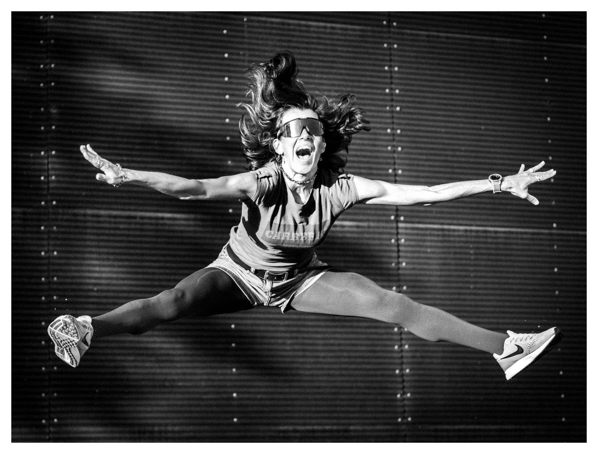 Black-and-white photo of a performer midair in a wide split jump, arms outstretched, wearing sunglasses, a T-shirt, shorts over leggings, and sneakers, with hair flying against a dark ribbed backdrop.