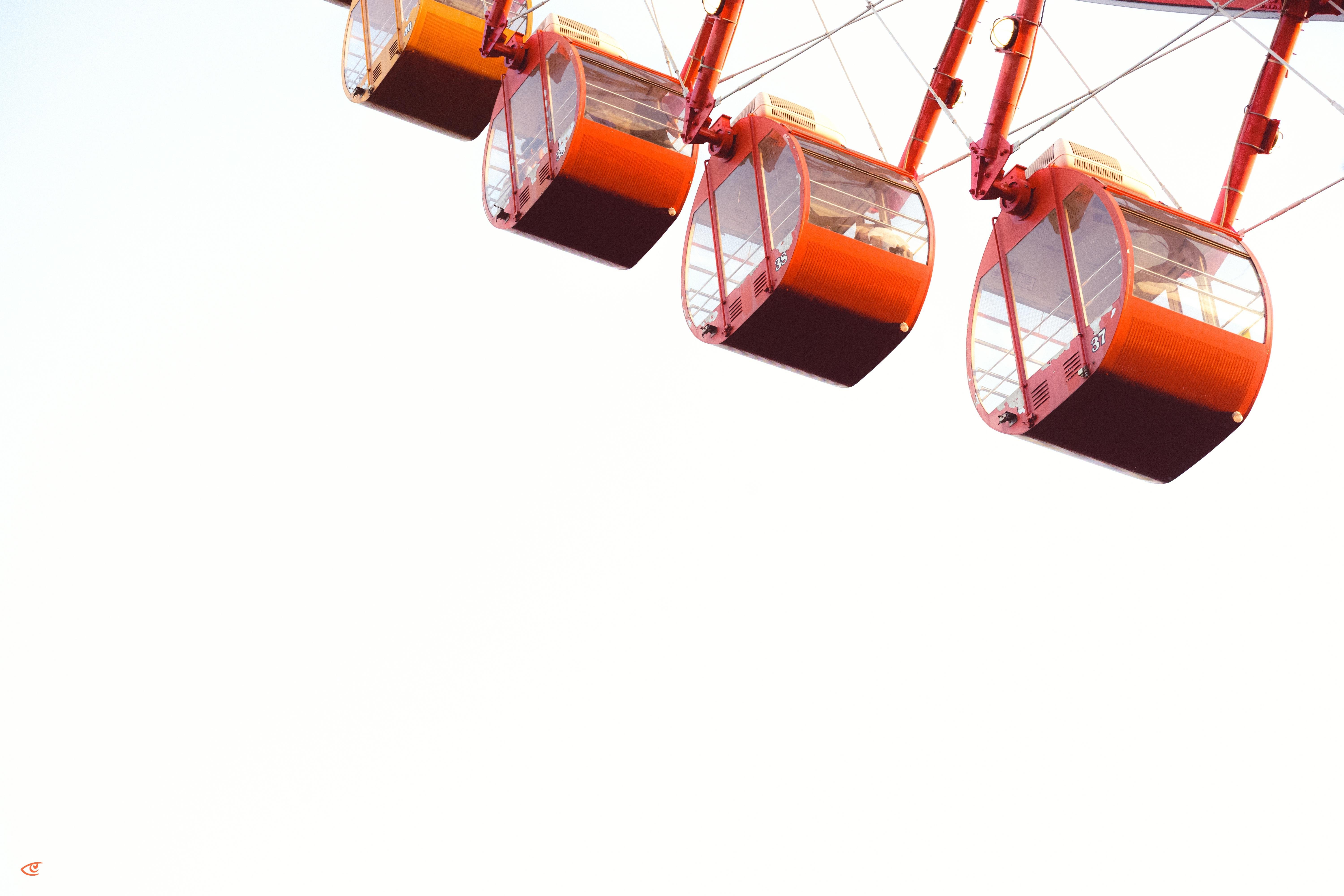 Four orange gondola cabins on a Ferris wheel hang diagonally across the upper-right of the frame against a blank white sky, with most of the image as negative space.