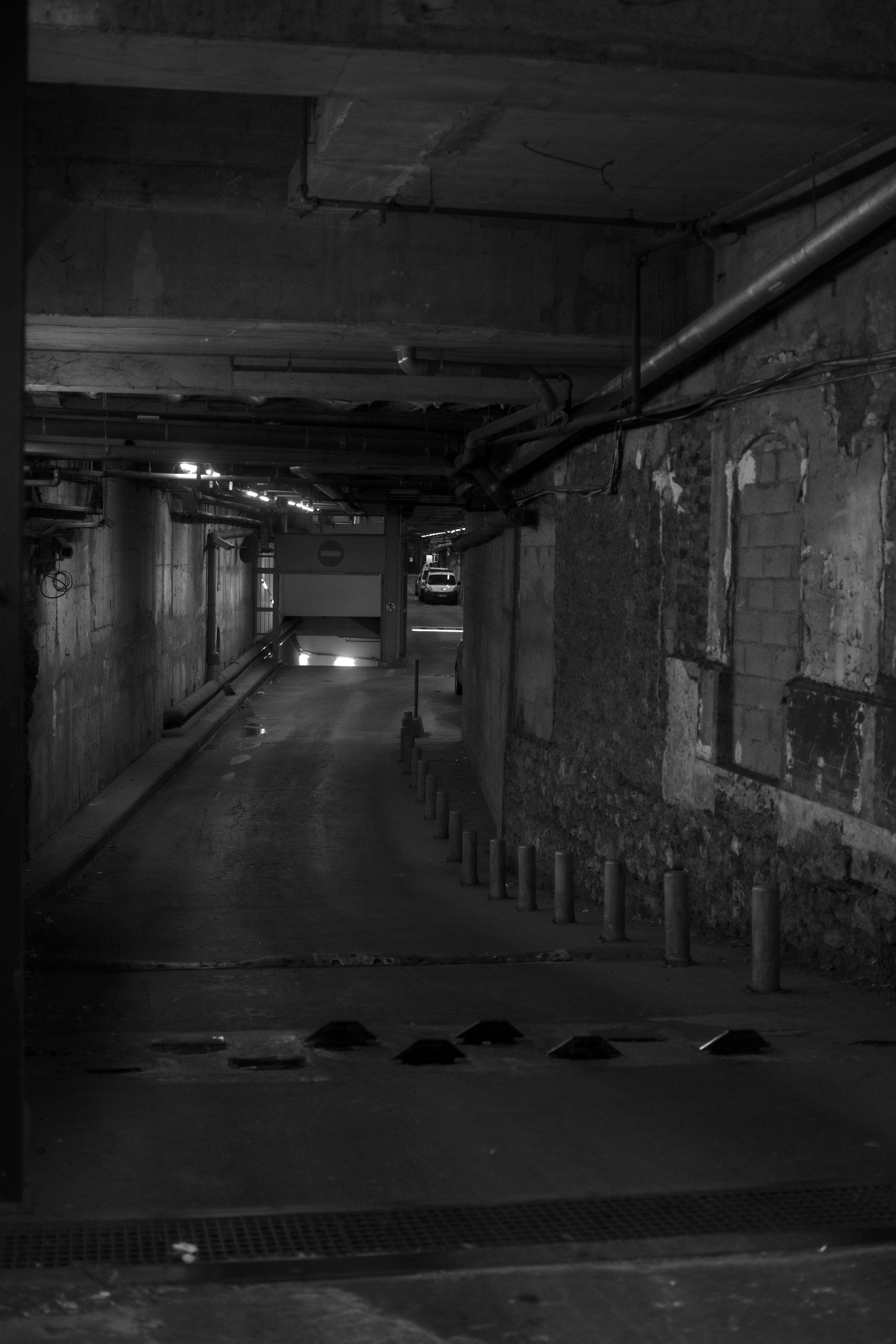 Photo format portrait, noir et blanc, de l’entrée d’un parking, le chemin légèrement courbé vers la droite. Les murs accusent leur âge et ne sont pas uniformes (briques, pierres, béton). Au premier plan, un ralentisseur formé de petites bosses en quinconces. À droite, des plots délimitent un chemin piéton. Au fond, le chemin se divise en deux et descend à gauche, et continue à droite, dans lequel on voit une voiture garée.