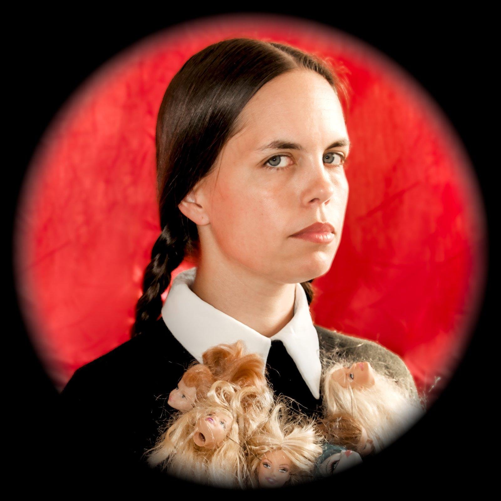 Une fille, cheveux sombres, attachés en 2 nattes, regarde la caméra d’un air strict voir lugubre, avec un collier fait de têtes de poupées type barbie, sur un fond rouge.