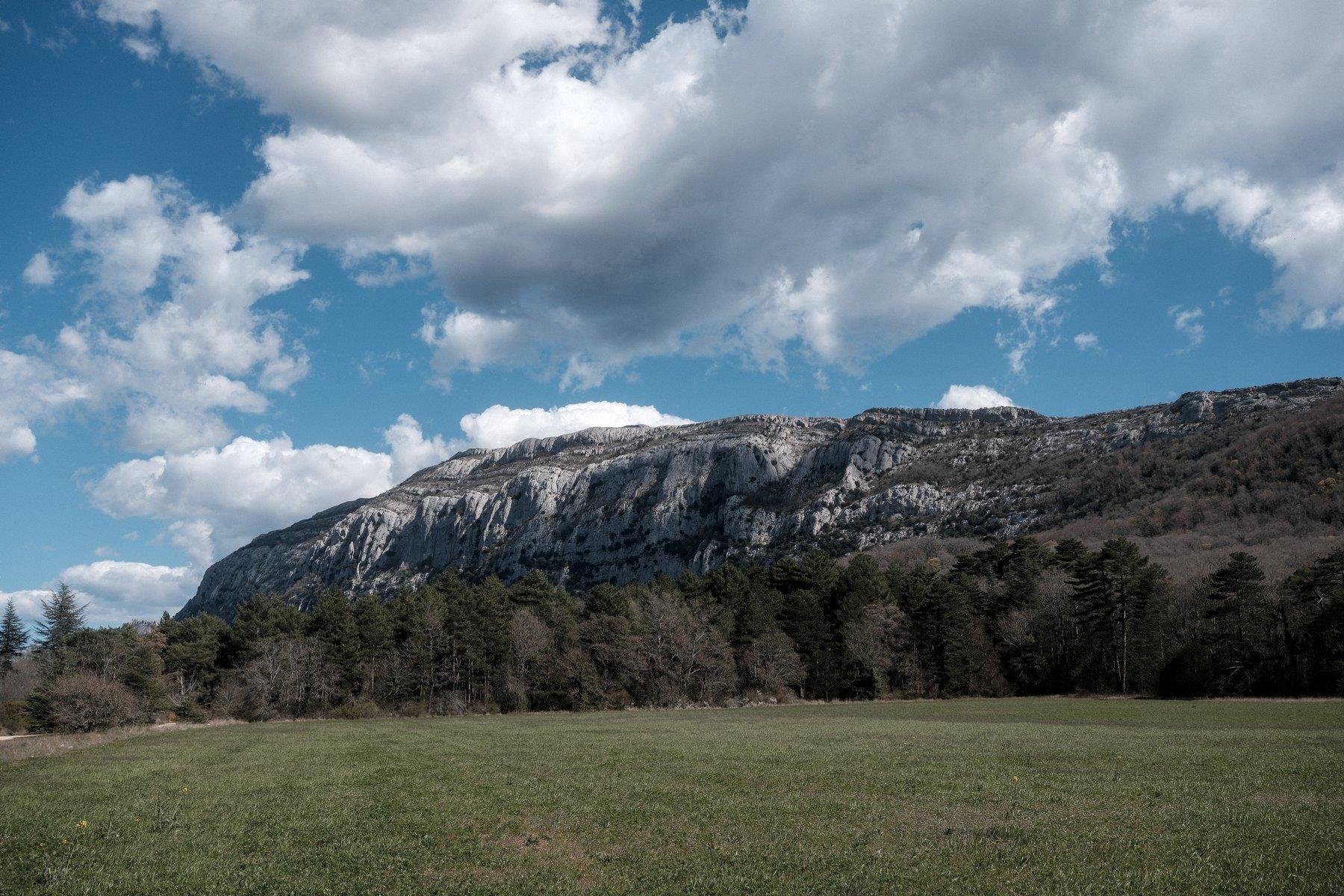 Photo horizontale. Paysage de la plaine verte, puis des arbres et enfin, le massif de la Saine-Baume, sous un ciel bleu peuplé de nuages.