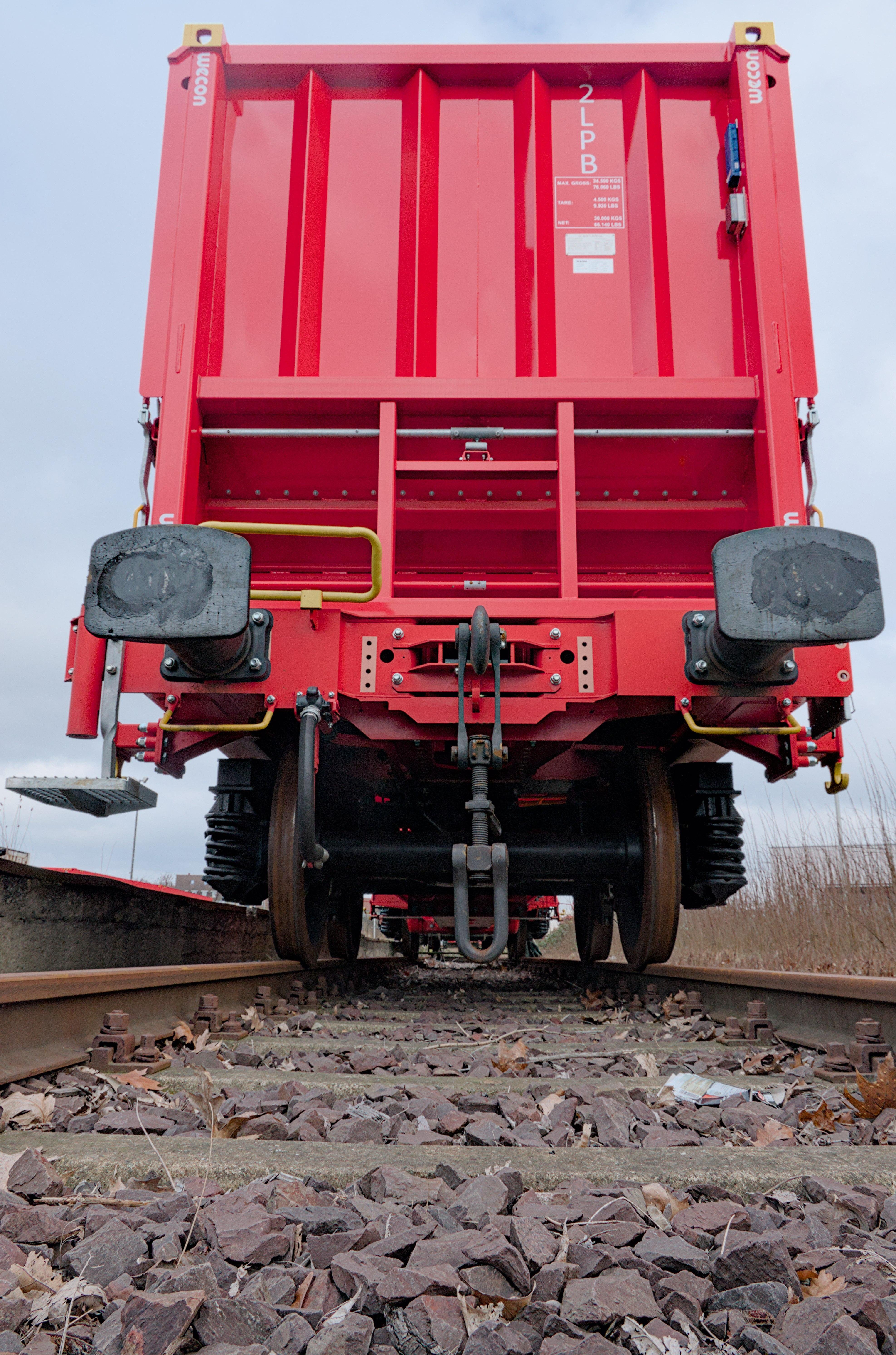 This image shows the end view of a red freight train car on a railway track. The photo captures the undercarriage and wheels, along with visible components like buffers and couplings. The track is visible with ballast (crushed stones) around it.