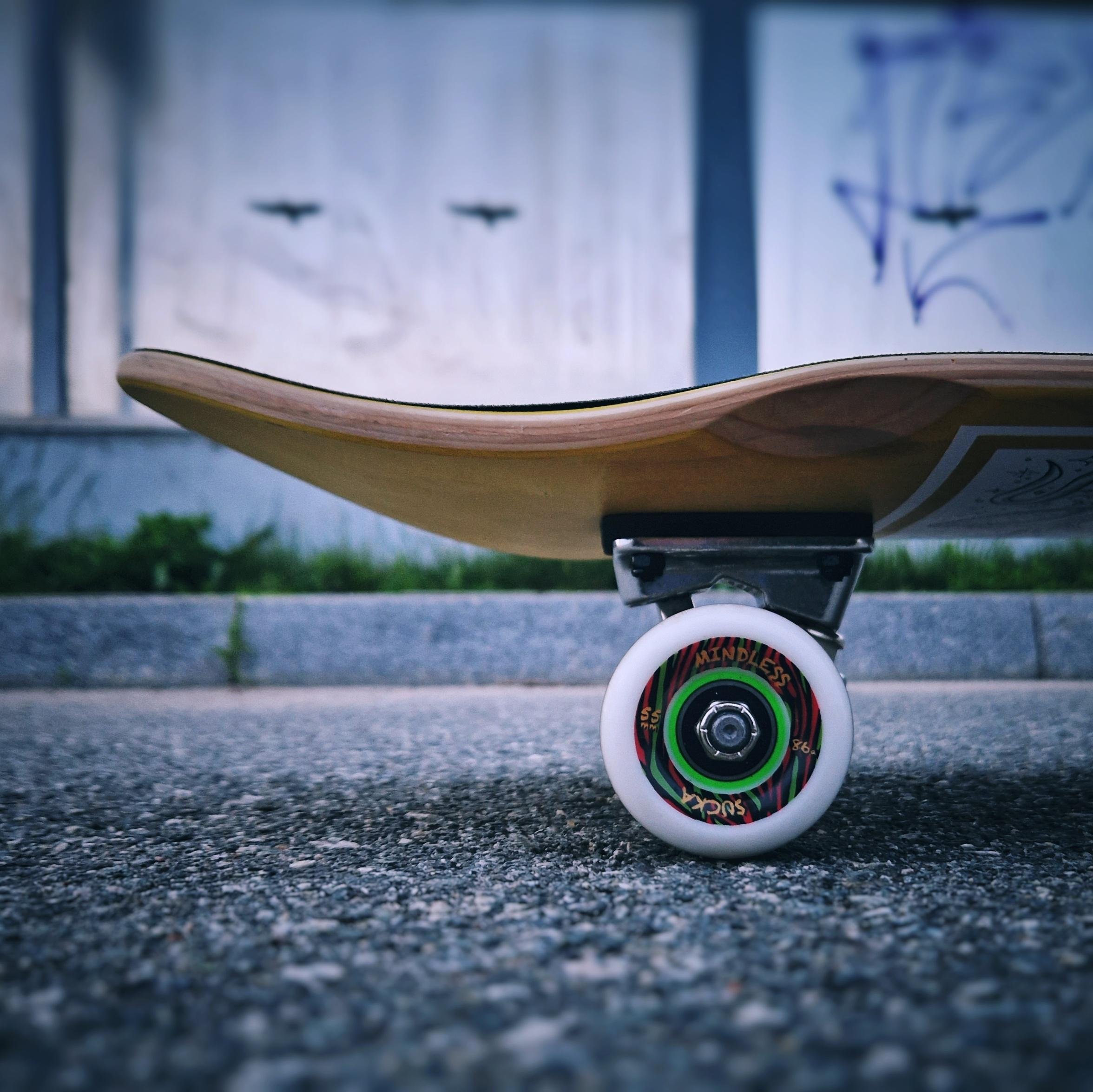 The image shows a close-up of a skateboard's wheel and part of its deck. The skateboard is on an outdoor surface, with the background slightly blurred, possibly showing a wall with graffiti. The focus is on the detailed design of the skateboard wheel.
