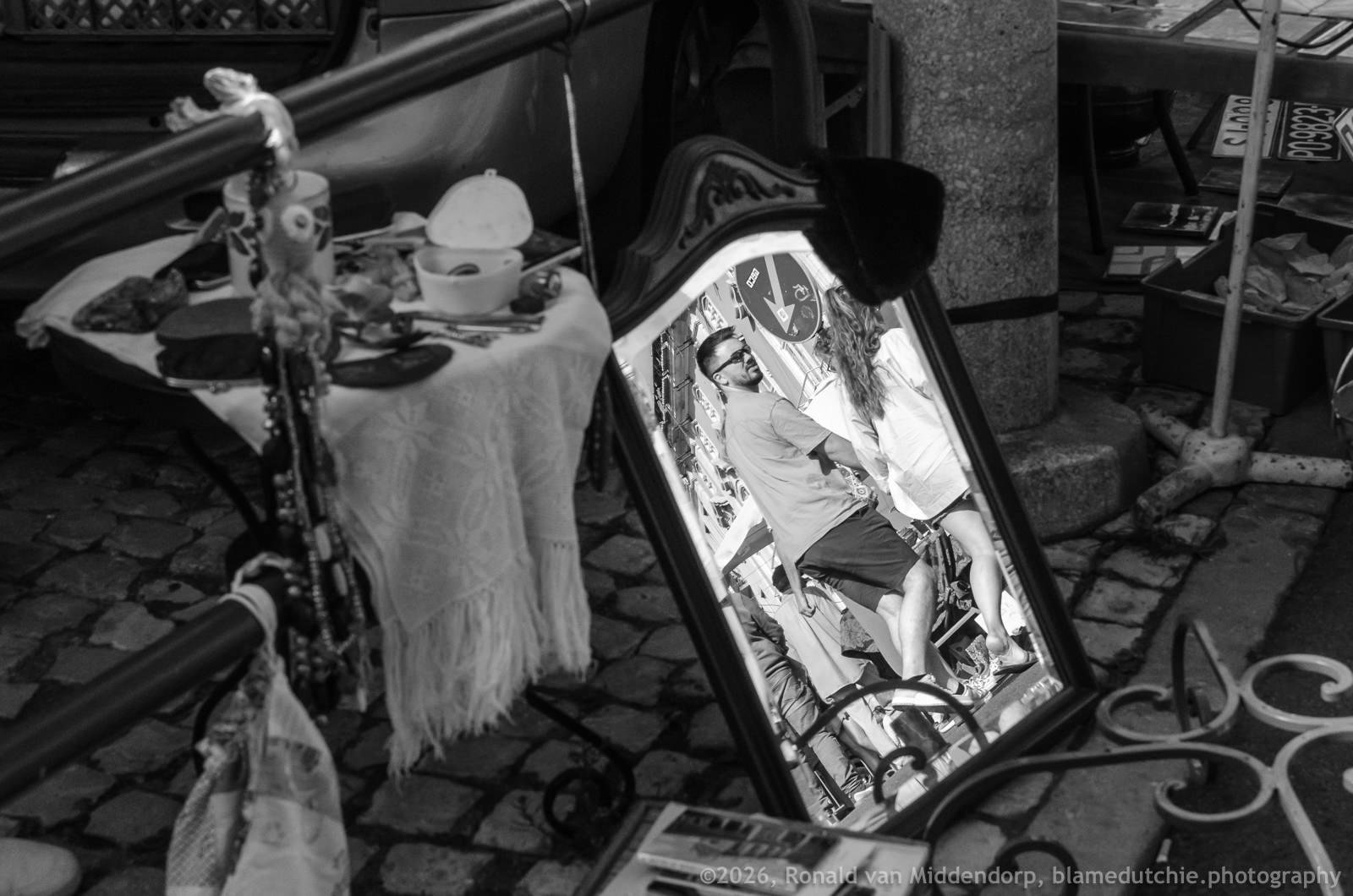Ornate framed mirror leaning on cobblestone ground at an outdoor market, reflecting two people standing close together; table with a fringed cloth, dishes, and hanging trinkets.