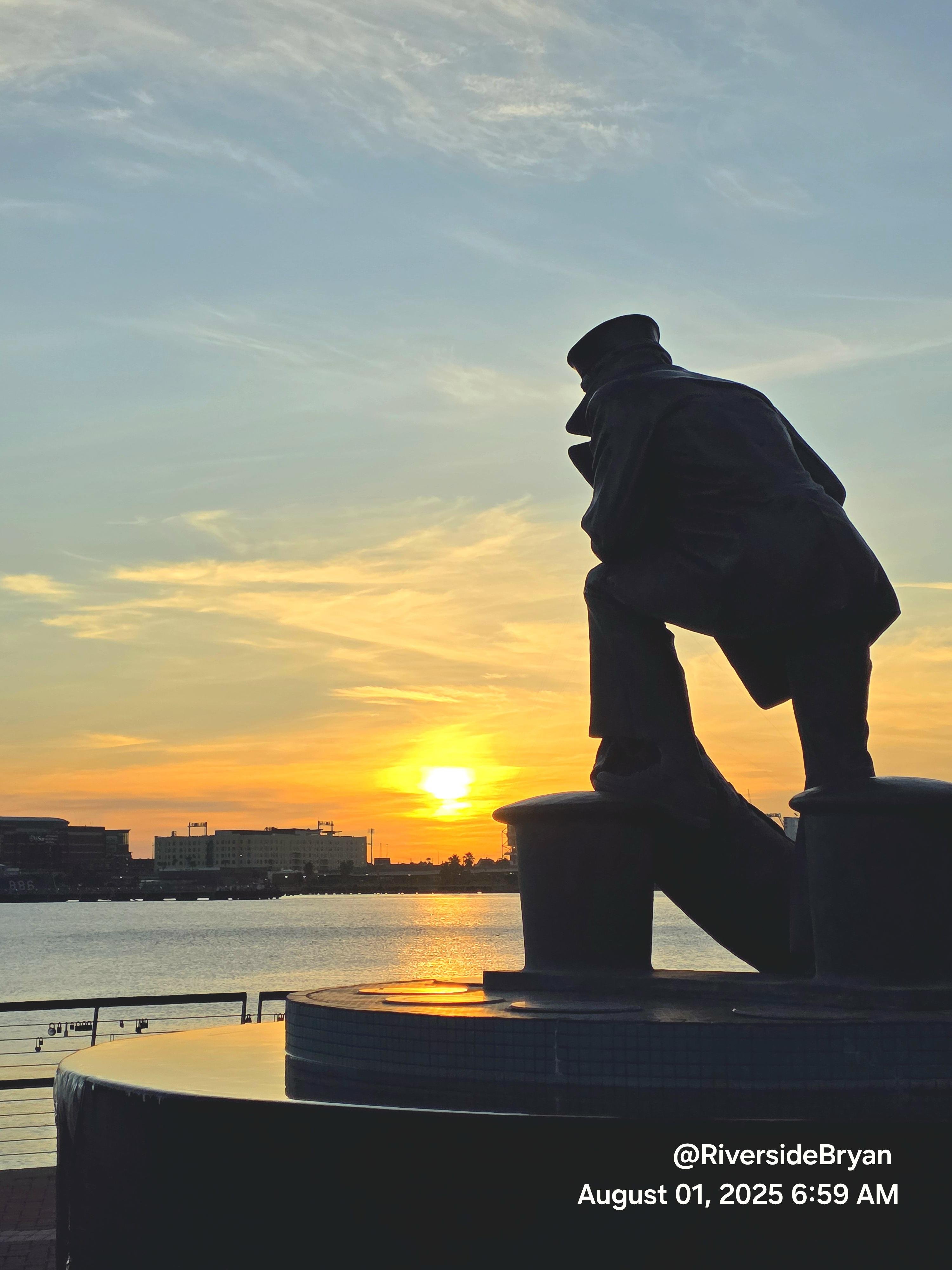 Silhouette of a statue depicting a sailor leaning on a ship's wheel, positioned against a sunrise with a partially cloudy sky. The sun is low on the horizon, casting a glow over the water and buildings in the background. The timestamp on the image reads August 01, 2025, 6:59 AM.