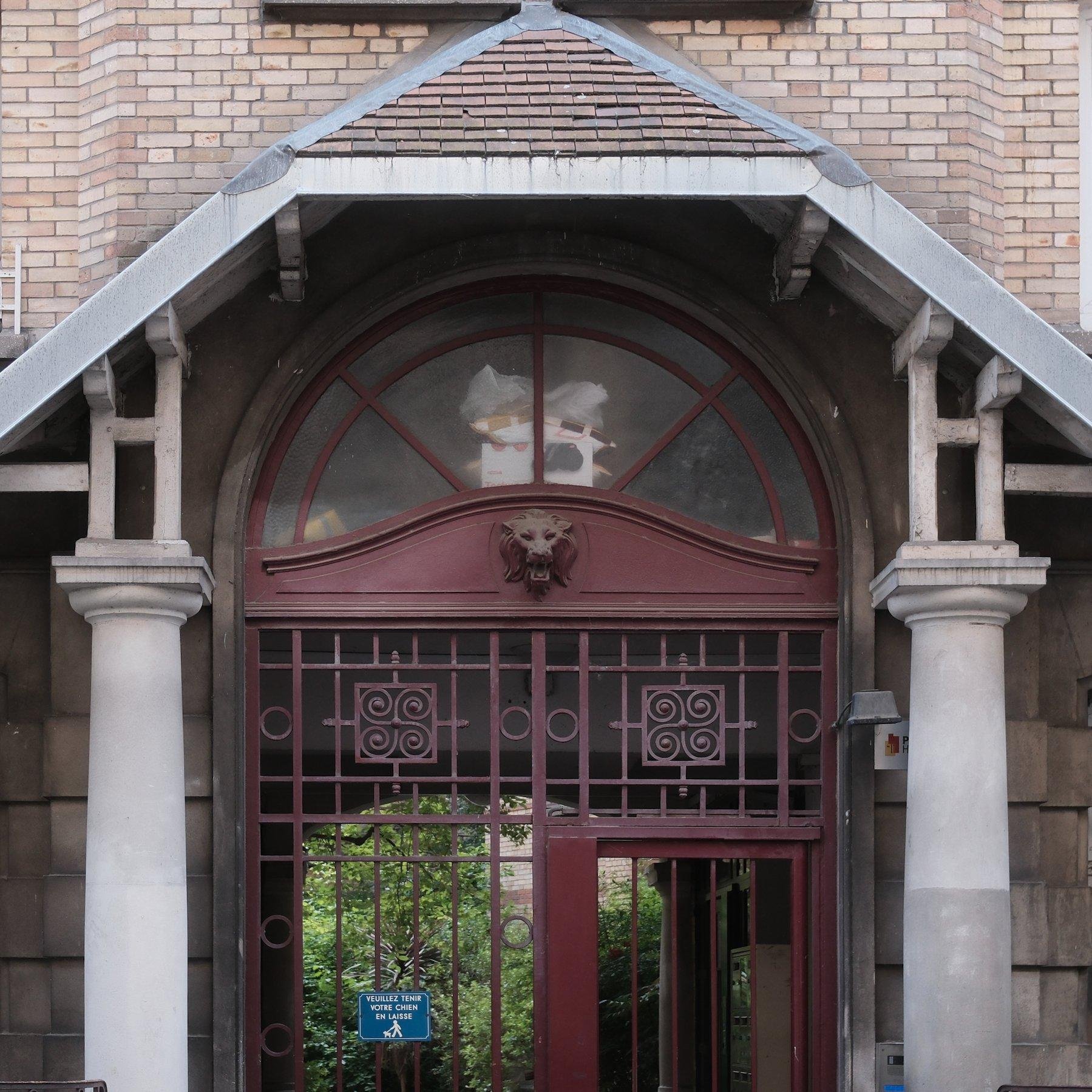 Photo carrée. La moité haute d’une porte d’immeuble ancien, en fer forgé peint en rouge, avec une tête de lion en décoration au dessus. Le tout se trouve sous un mini toit tenu par 2 colonnes