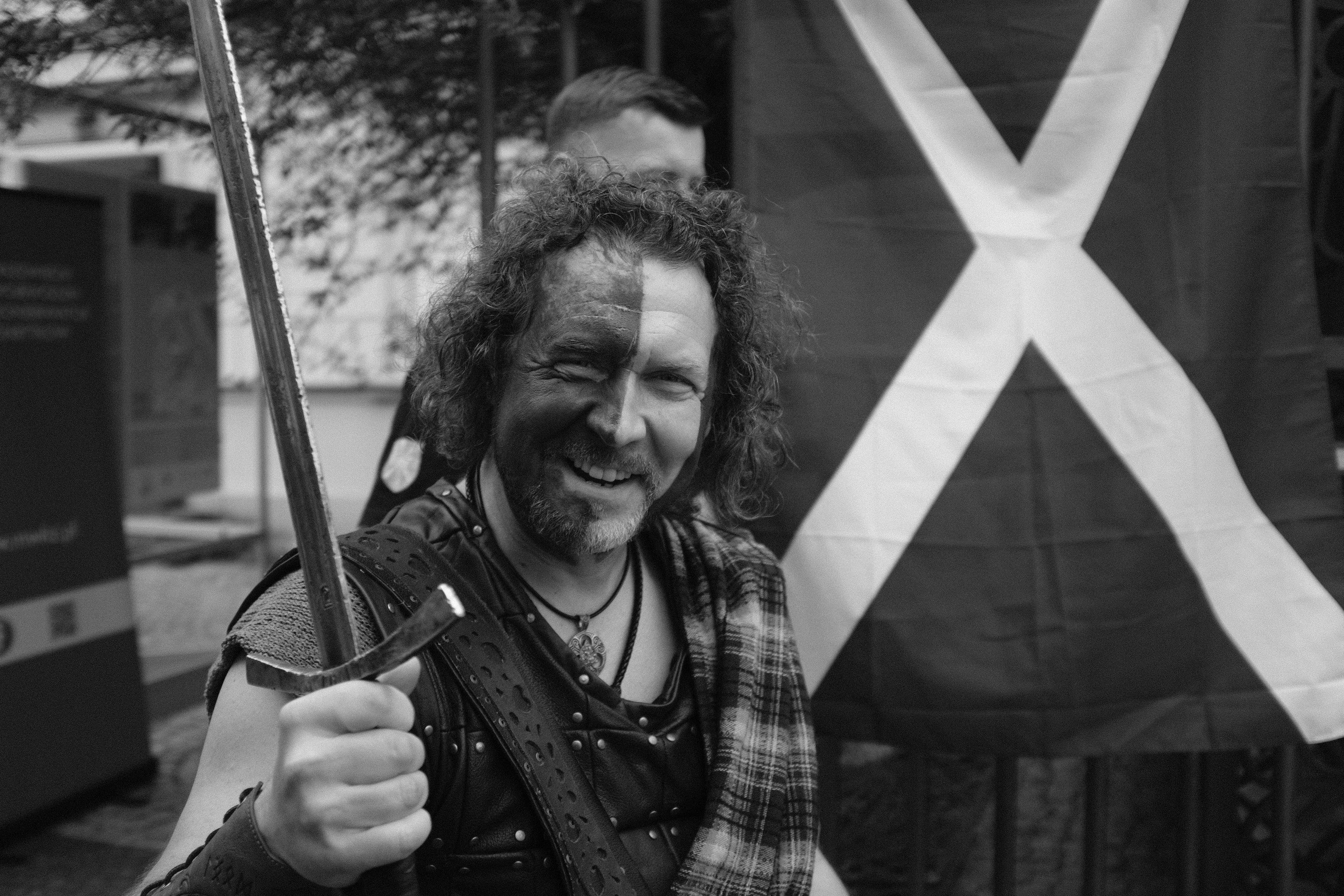 Man dressed in medieval warrior attire with face paint, holding a sword, and wearing a plaid sash. Scottish flag in the background. Black and white photograph with another person partially visible behind him.