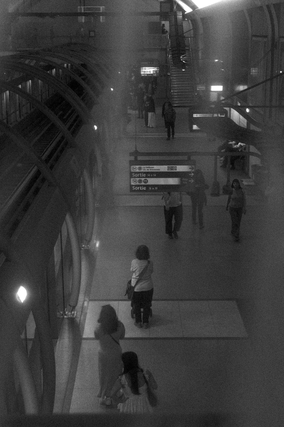 Photo verticale en noir et blanc. Vue de dessus sur un quai de métro avec portes automatiques. Une vieille dame pousse son chariot de couses, seule au milieu des passants