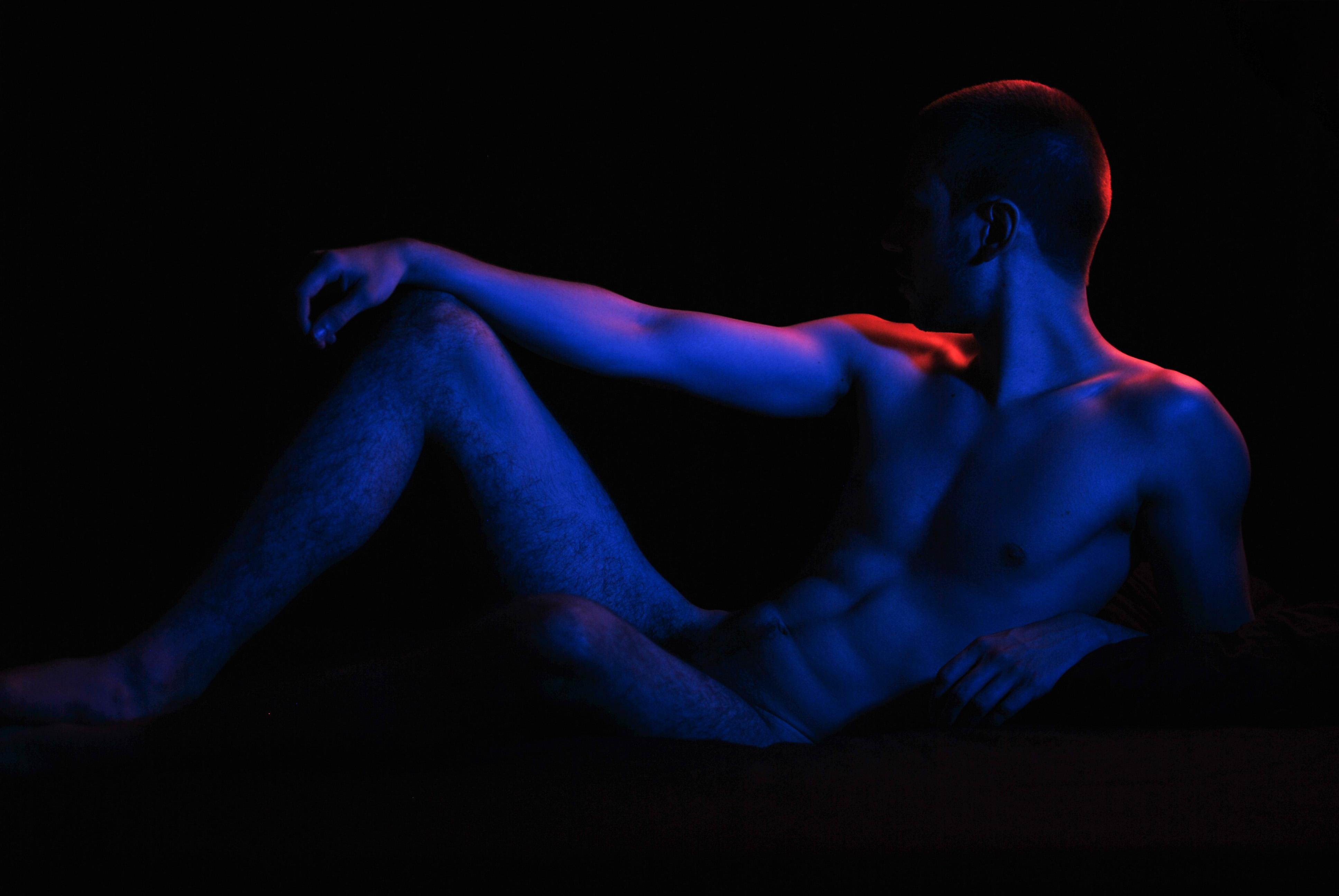 This image shows a silhouette of a male figure reclining in a dramatic pose. The lighting is low-key with blue and red tones highlighting the contours of his body, creating a moody and artistic effect. The background is dark, emphasizing the contrast between light and shadow.