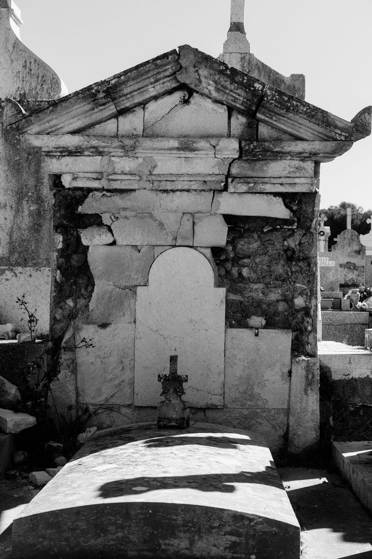 Photo verticale en noir et blanc. Une tombe de face, dont la grosse stèle est entrain de se craqueler sous l’effet de l’érosion