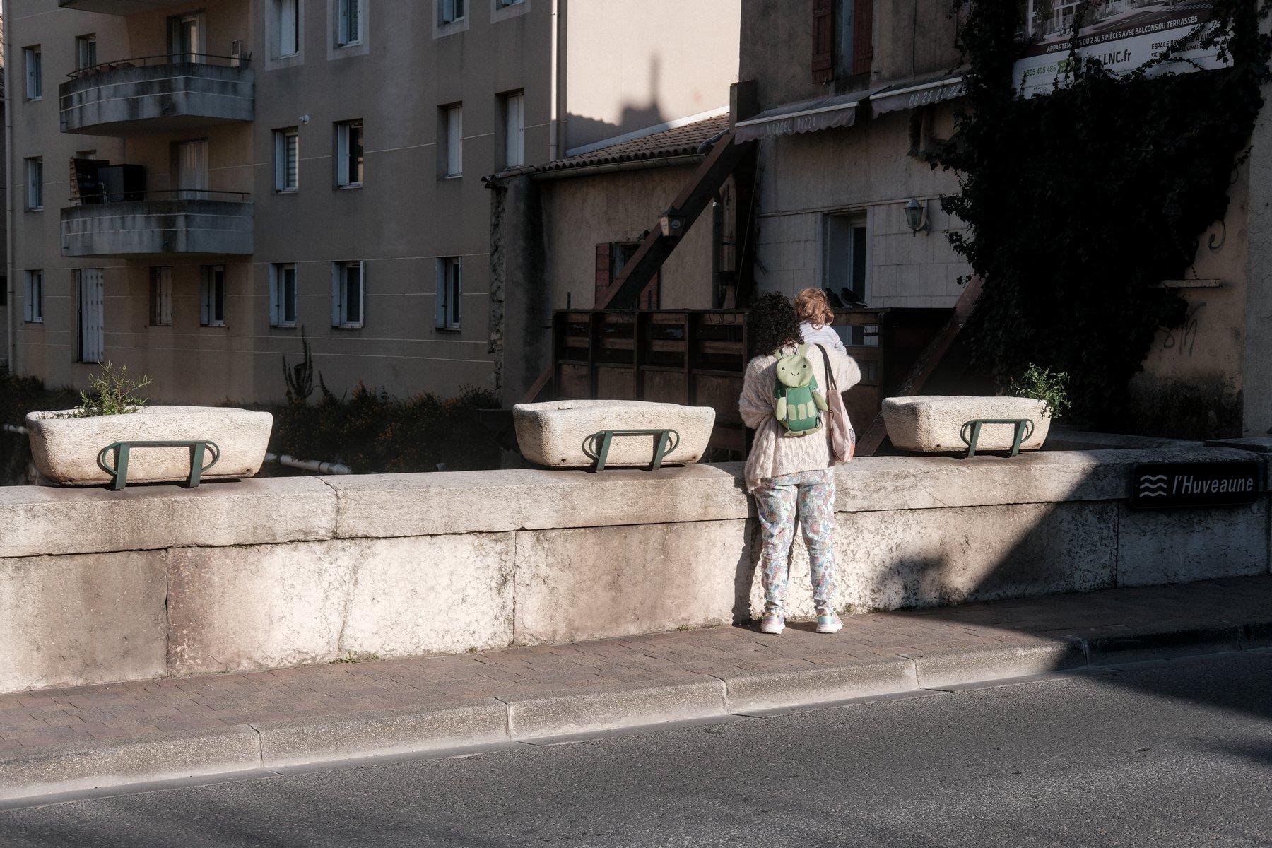 Photo horizontale. Une femme tiens fermement une fillette sur le muret de sécurité d’un pont