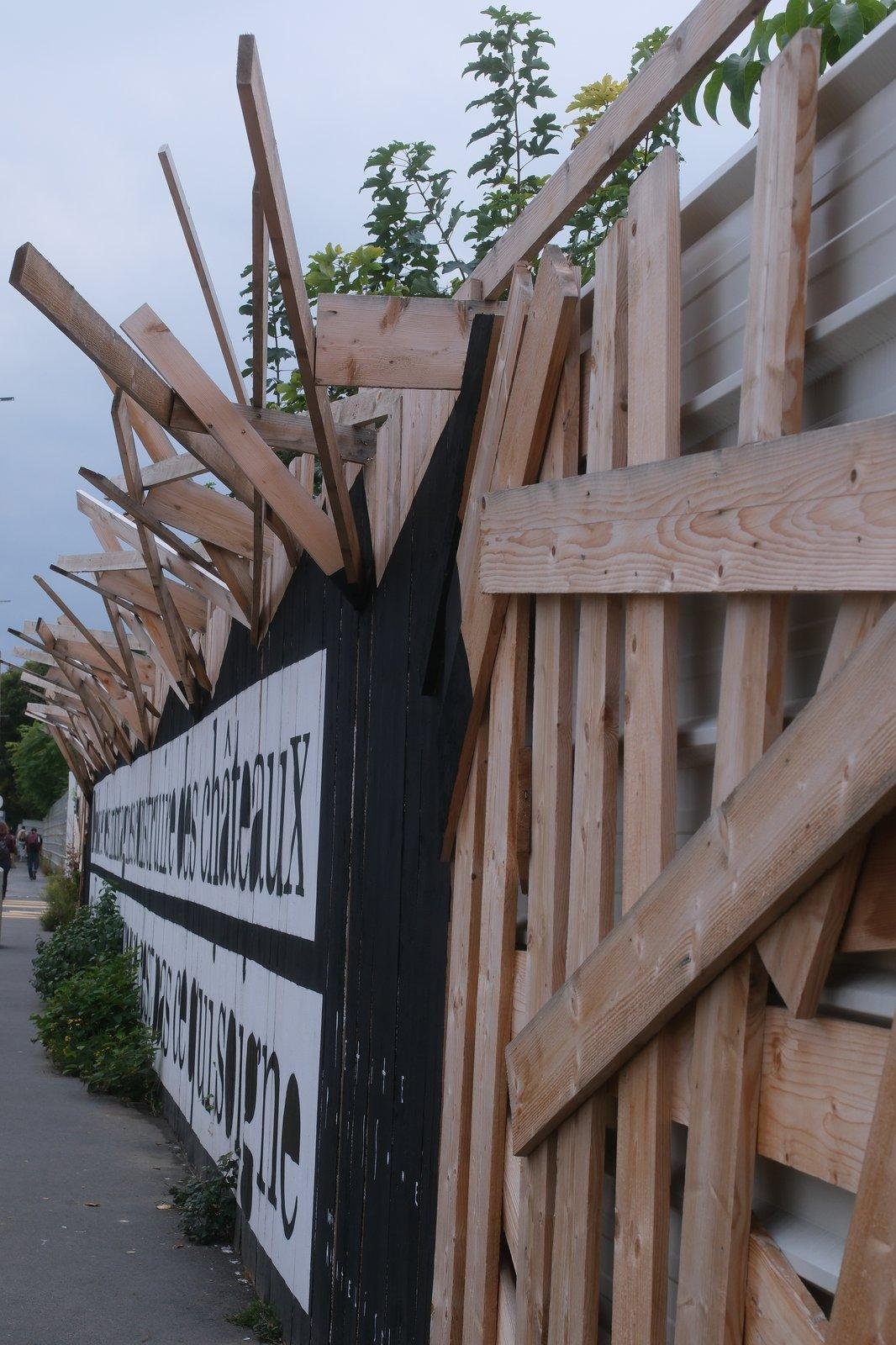 Photo verticale. Un mur protégeant un chantier, avec des planches de bois mises un peu en vrac au premier plan, et ensuite, mises comme pour faire des piques de défense/barbelés en bois