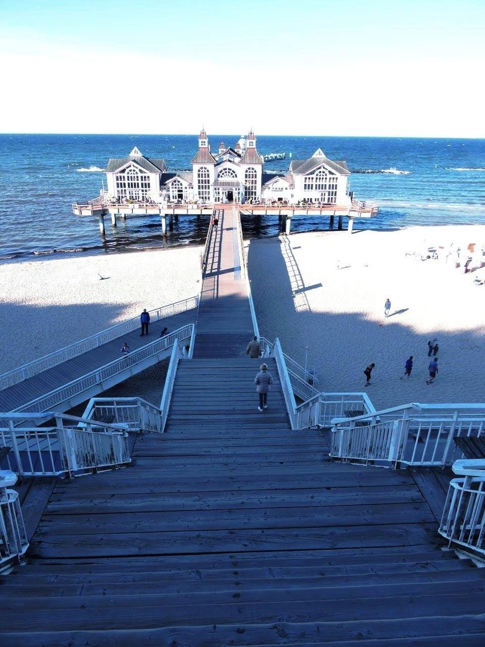 Wide wooden staircase leading down to a sandy beach with a pier extending into the ocean. At the end of the pier, a large pavilion with intricate architectural details sits above the water. People are visible walking along the stairway, beach, and pier. The ocean appears calm under a clear blue sky.