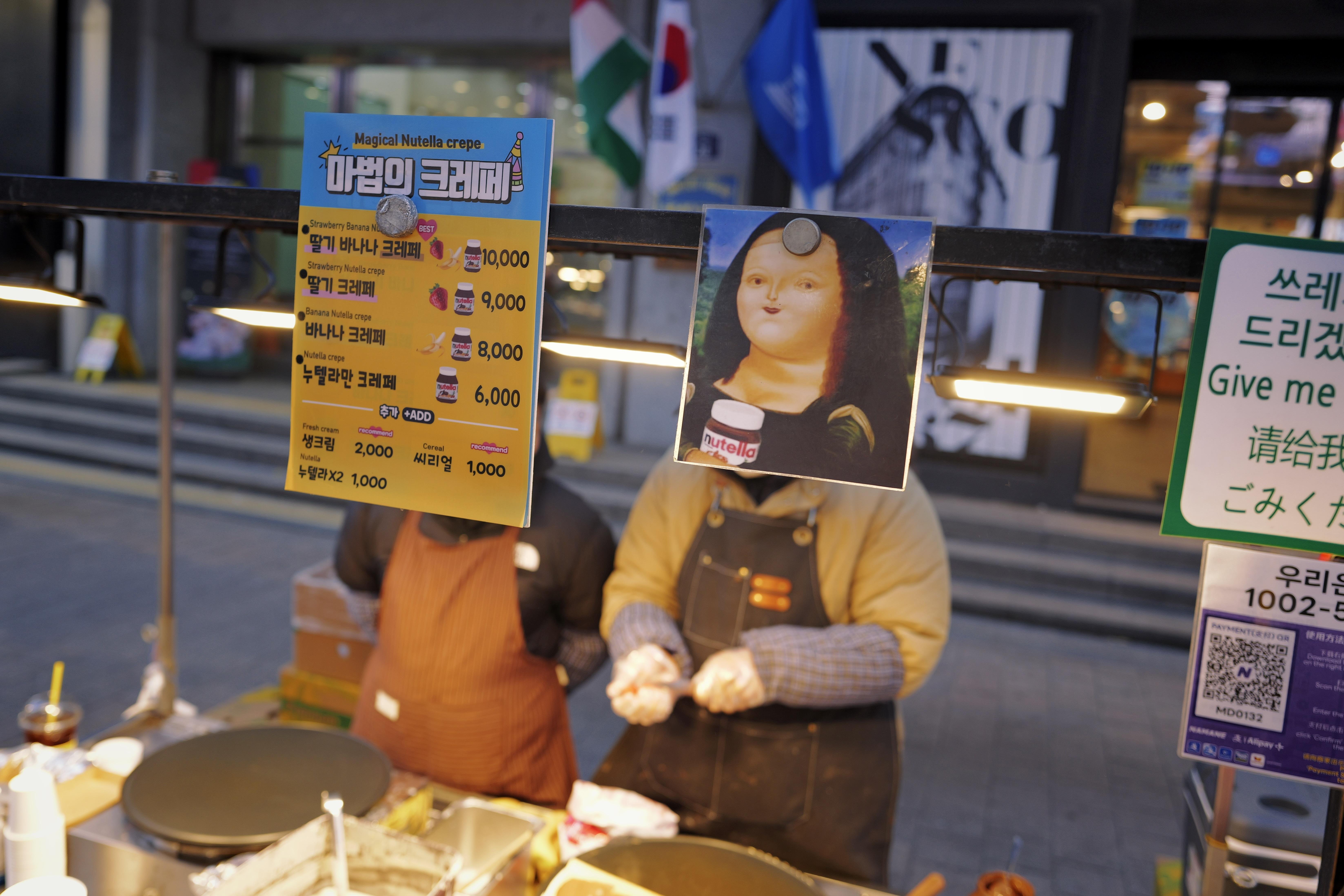This image shows a street food stall where crepes are being prepared. There are menus displayed in multiple languages, including Korean and English, listing various crepe options with prices. One of the signs humorously features a parody of the Mona Lisa holding a jar of Nutella. The stall is located outdoors, with flags and signs in the background.