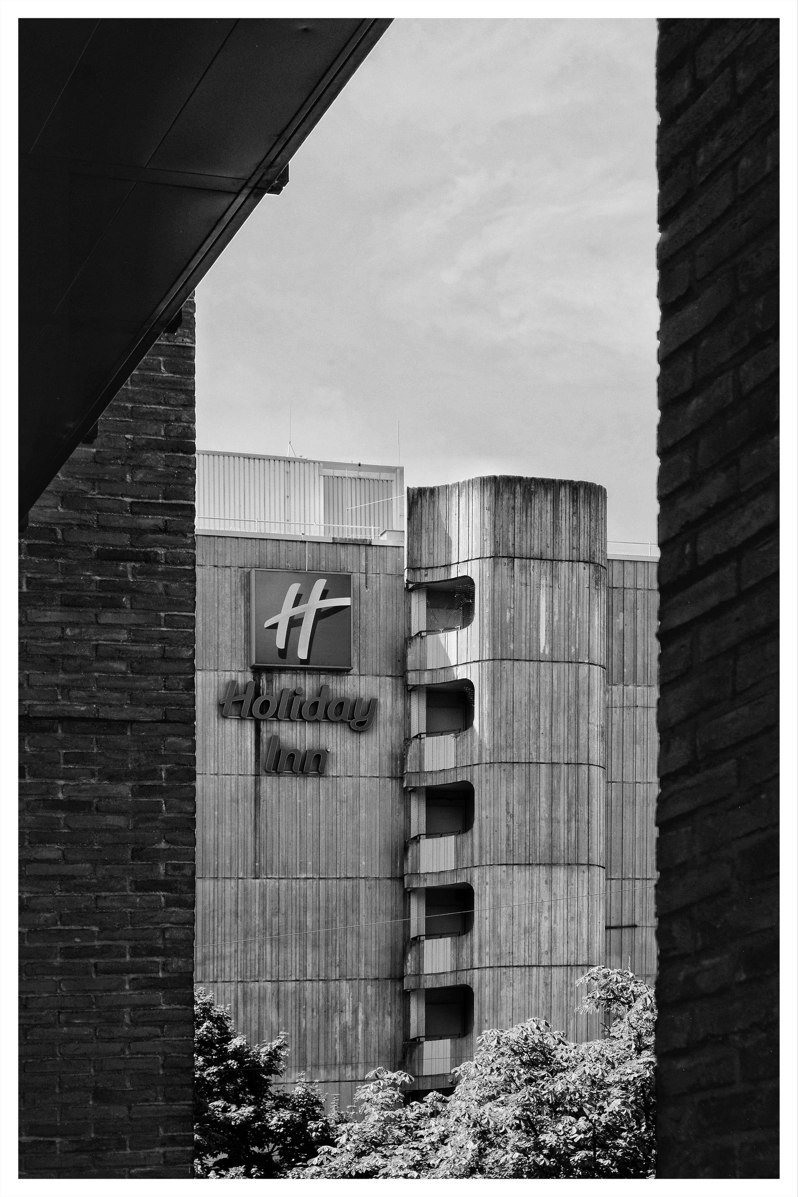 Black-and-white photo of a Holiday Inn building with a vertical strip of recessed windows, framed by dark brick walls on the left and right and an overhanging roof at the top left, with trees along the bottom edge.