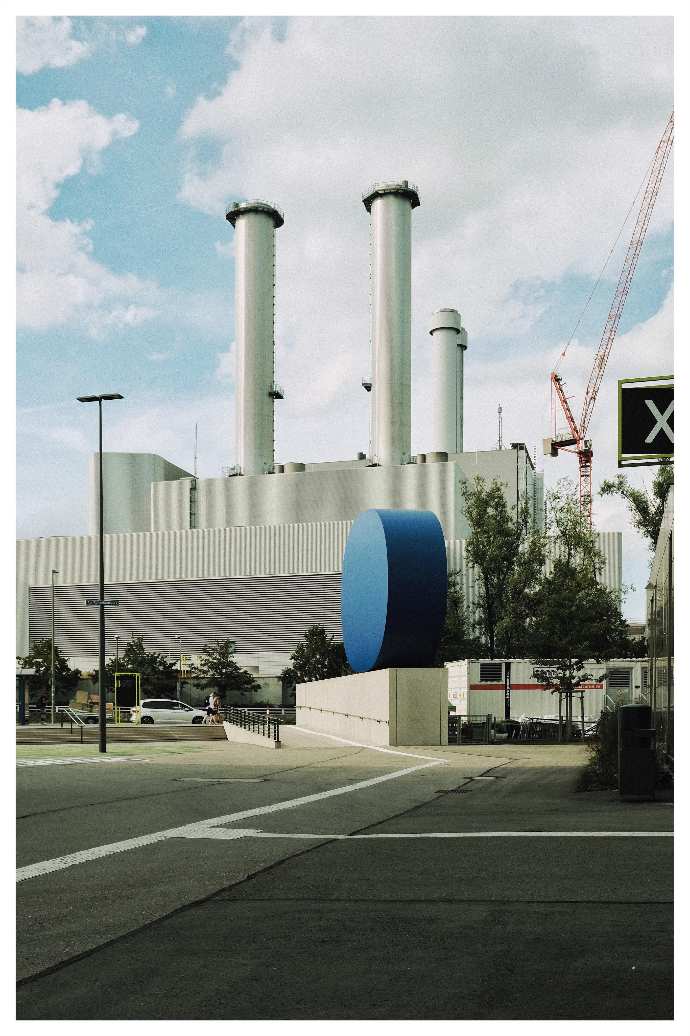Industrial facility with three tall cylindrical smokestacks behind a low gray building; a large blue cylindrical structure sits on a concrete base in the foreground near a road, with trees, parked cars, a streetlight, and a construction crane under a cloudy sky.