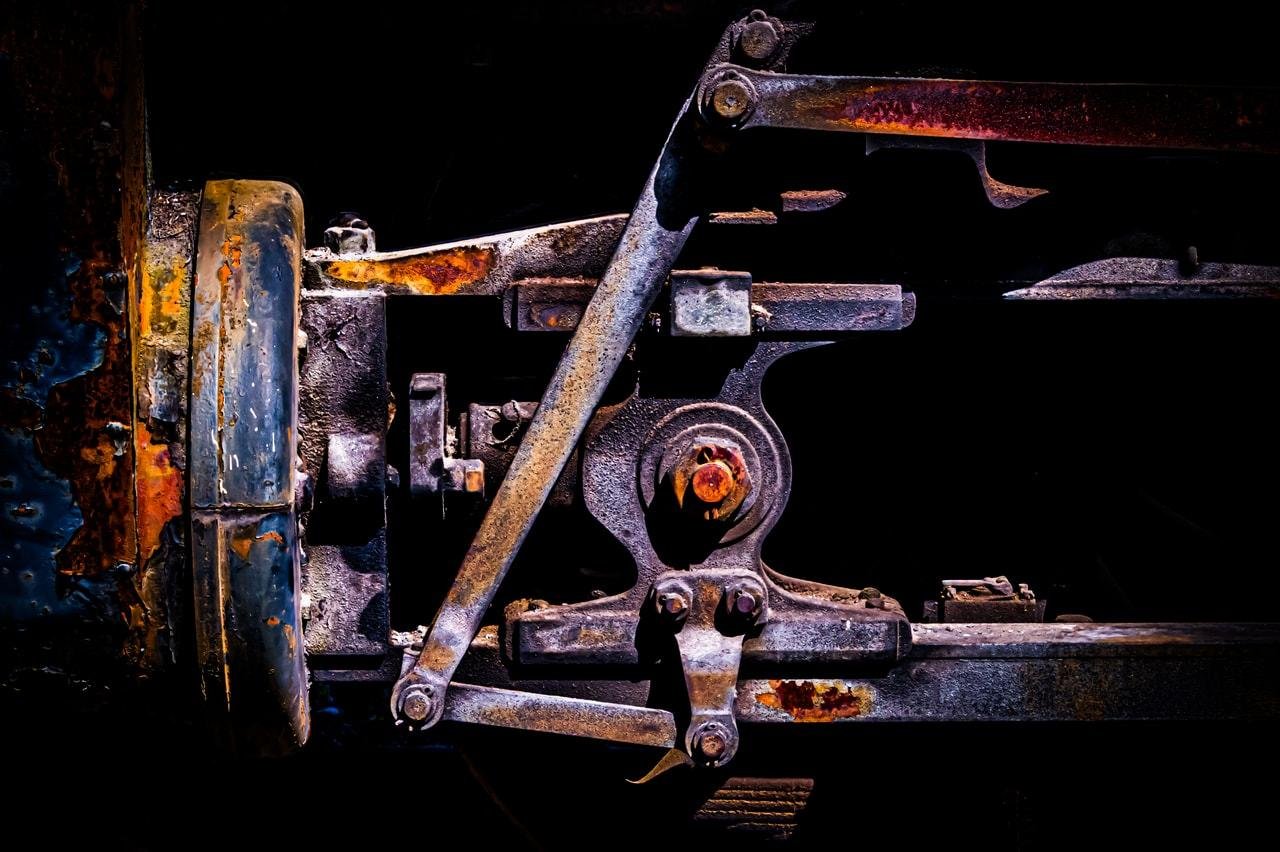 Close-up of a mechanical assembly with visible rust and wear, featuring interlocking metal components and a wheel on the left. The machinery has a mixture of dark, oxidized, and rusted metal textures, with bolts and joints prominently displayed. The background is dark, emphasizing the industrial elements.