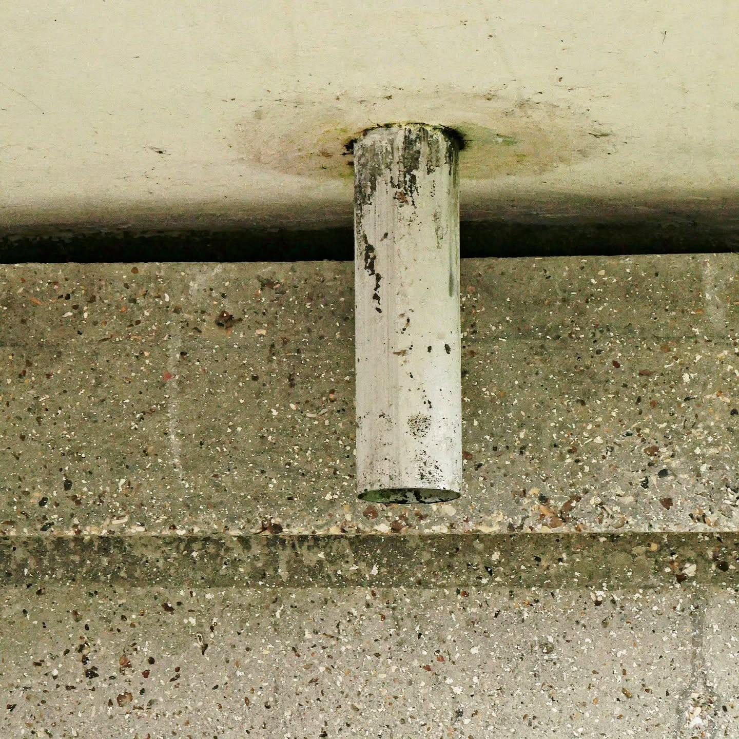 This image shows a white cylindrical white painted metal object, mounted against a textured concrete surface. The tube appears to have some wear on it, and it is projecting out from the surface.