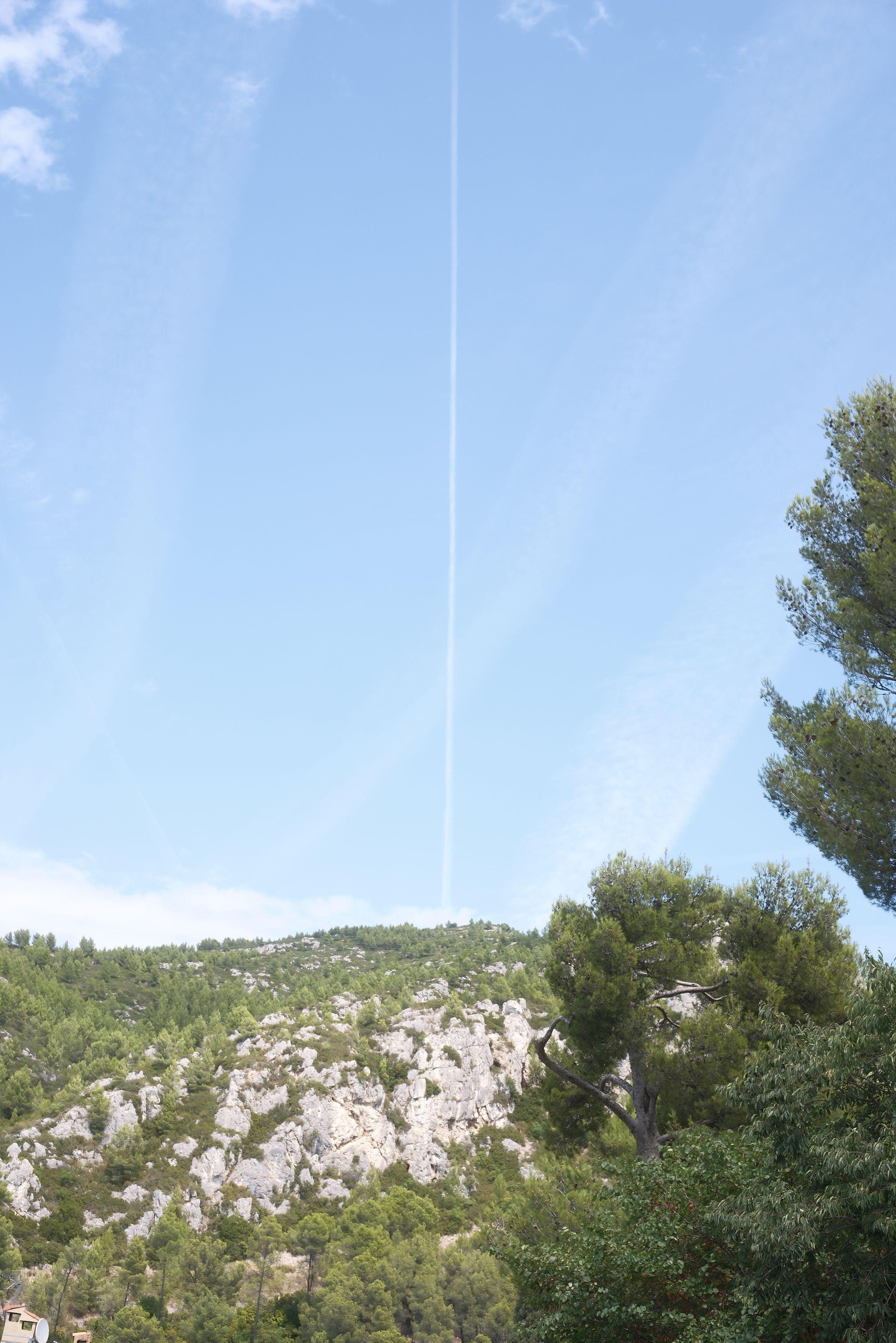Photo verticale. En bas de la photo, les montagnes de provence, puis le ciel bleu avec un trait droit vertical blanc dans le ciel, coupant l’image en 2