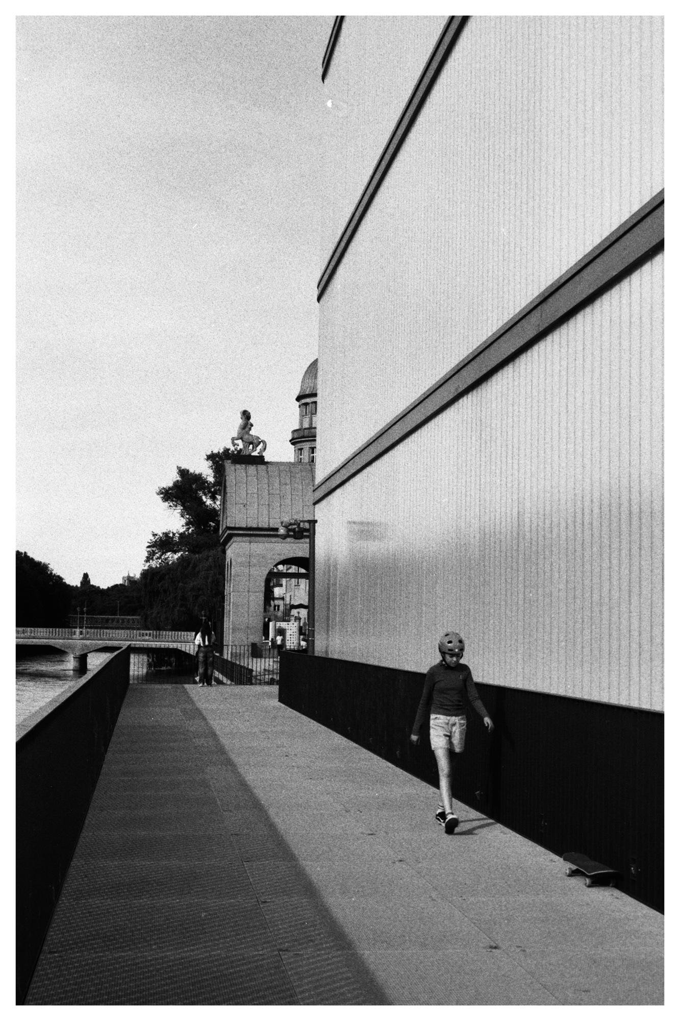 Black-and-white photo of a riverside walkway beside a tall building wall; a child wearing a helmet walks near a skateboard in the foreground, with a bridge and distant buildings visible in the background.