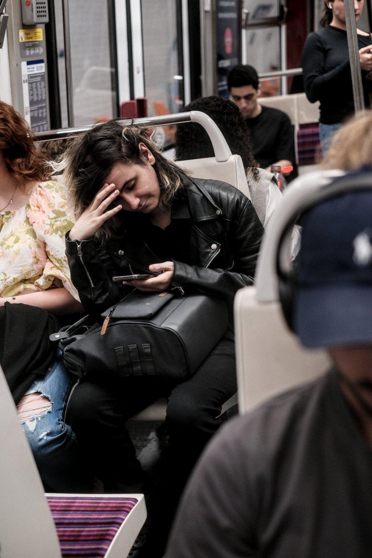 Photo verticale. Un homme assis dans un carré pour 4 dans le métro, cheveux longs avec les pointes teintes. Il porte un blouson noir en cuir, jean noir. Sac à dos sur ses genoux, il consulte son téléphone le bras posé sur le sac et la main droite se tenant le front